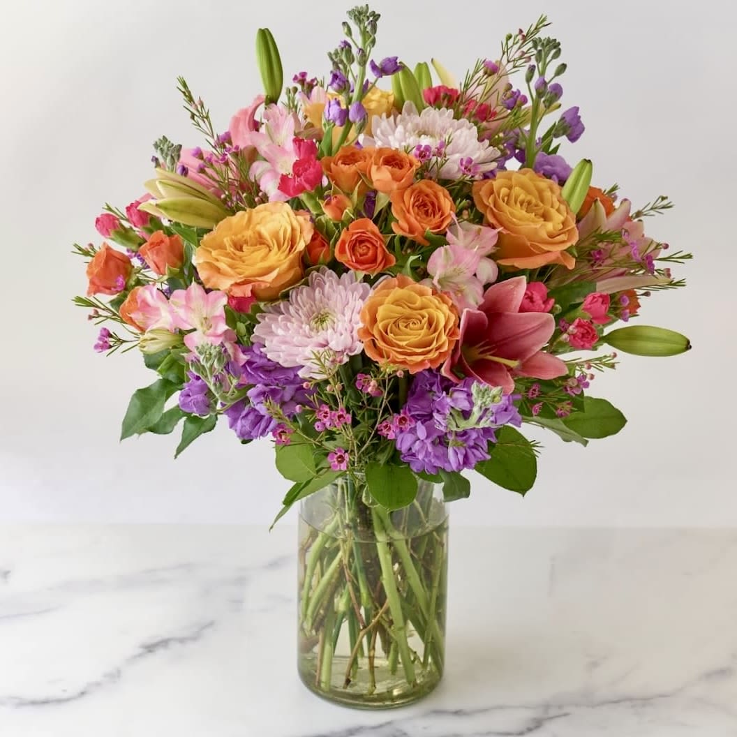 Bright mixed bouquet of orange roses, pink lilies, and purple blooms in a clear glass vase