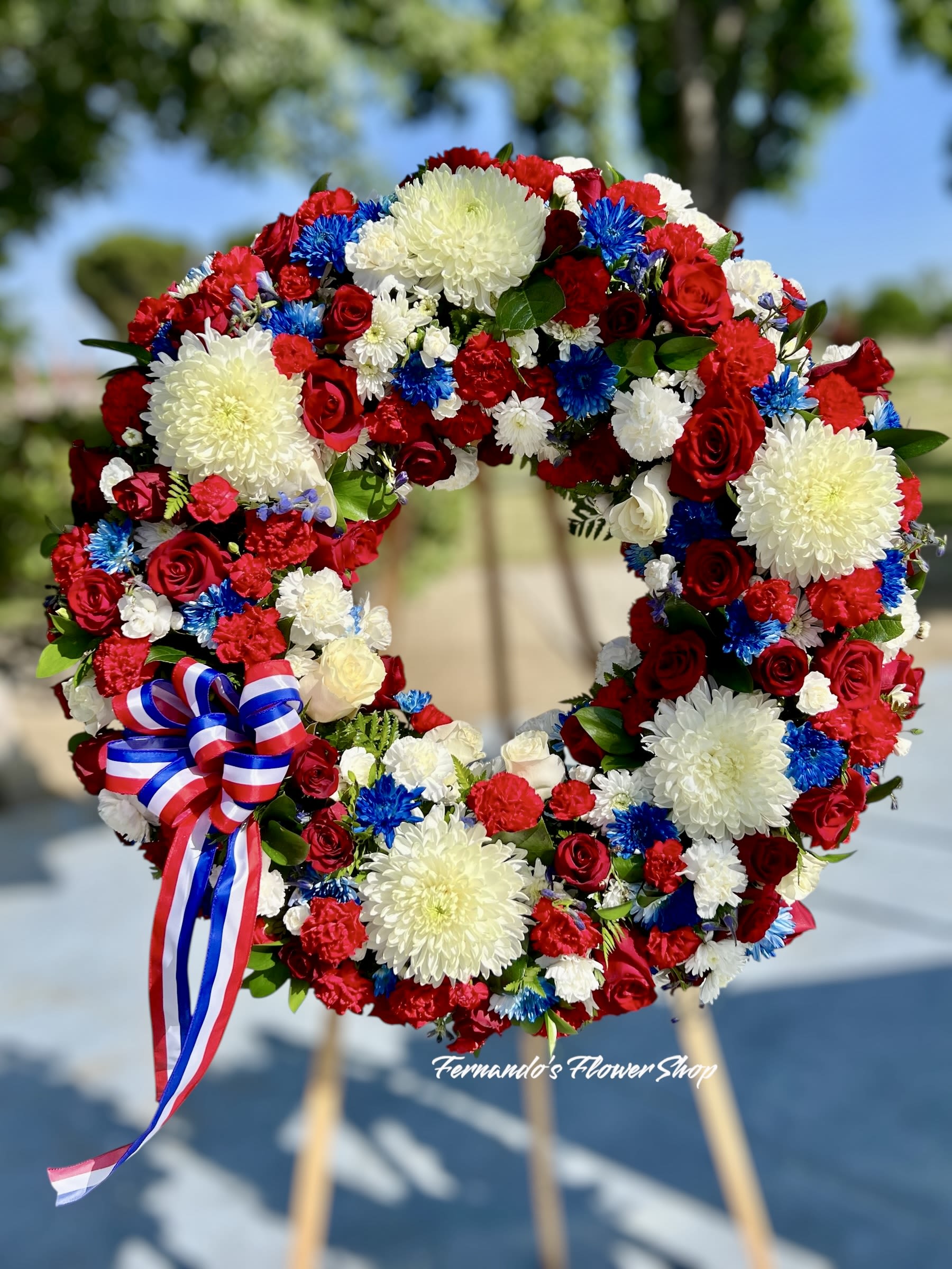 Honor &amp; Pride - A patriotic wreath that honors with pride and love. This vibrant tribute features bold red roses, white chrysanthemums, and striking blue accents, finished with a red, white, and blue ribbon. A beautiful way to show respect and remembrance for a hero.