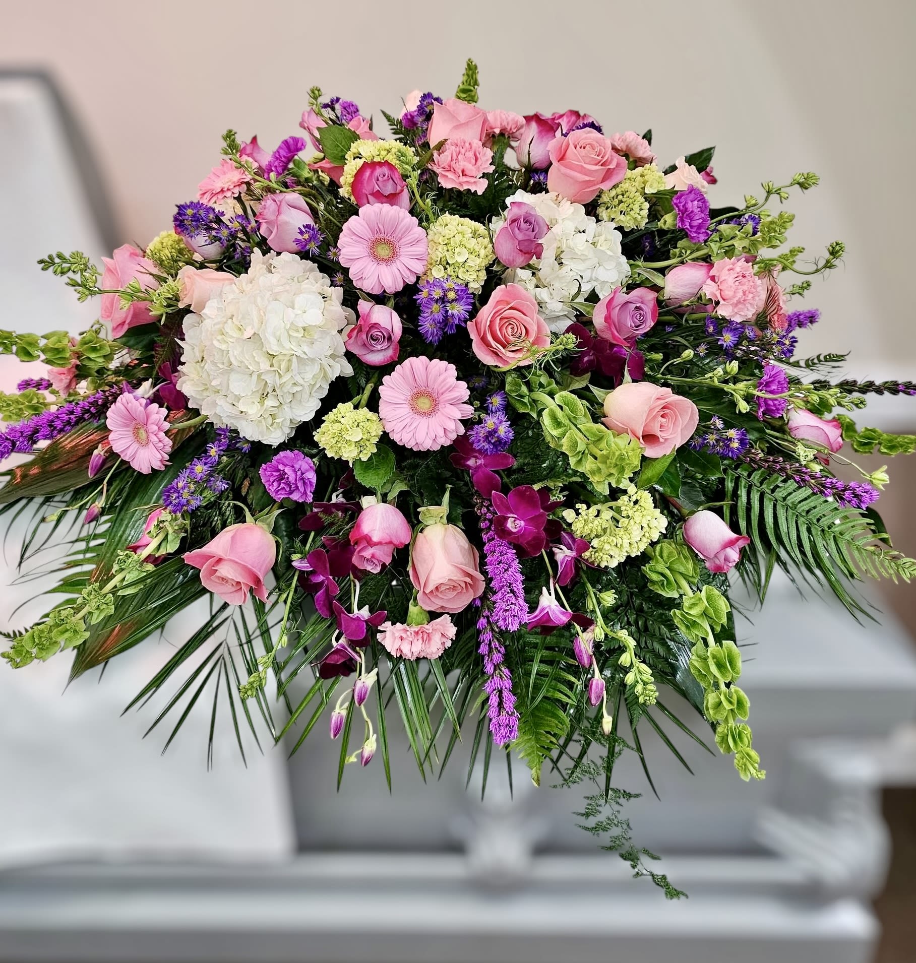 Pink and purple casket spray with roses, gerbera daisies, orchids, and hydrangeas on a white casket.
