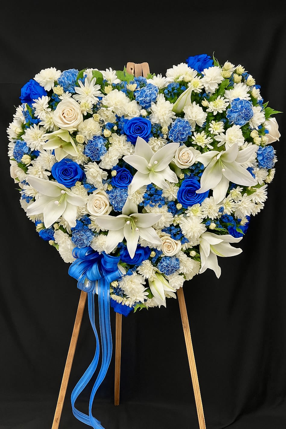 Heart-shaped blue and white standing floral wreath with roses, lilies, and carnations on a wooden easel.