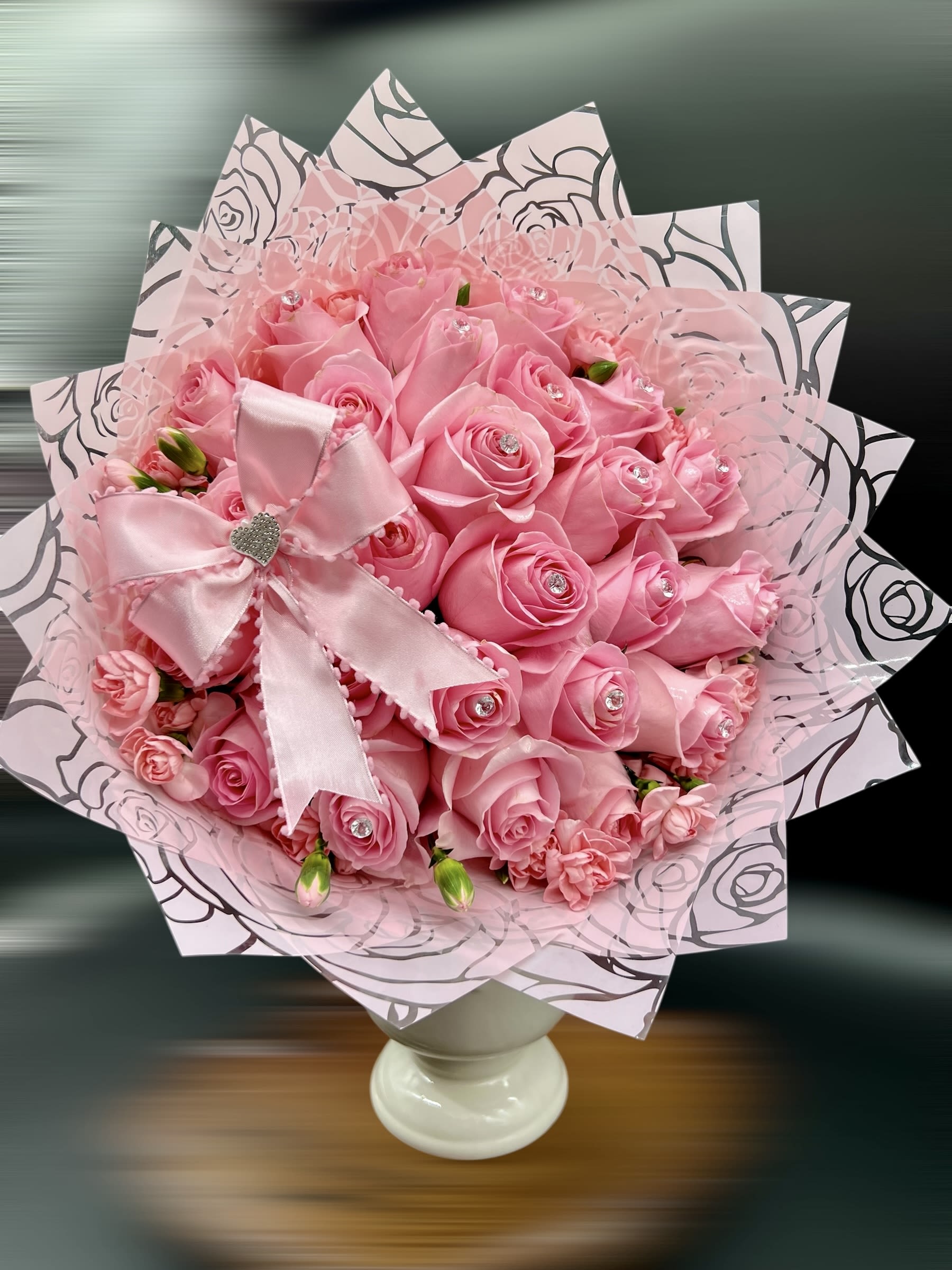 Hand-tied bouquet of pink roses with jeweled centers in pink wrap and bow on a white pedestal vase