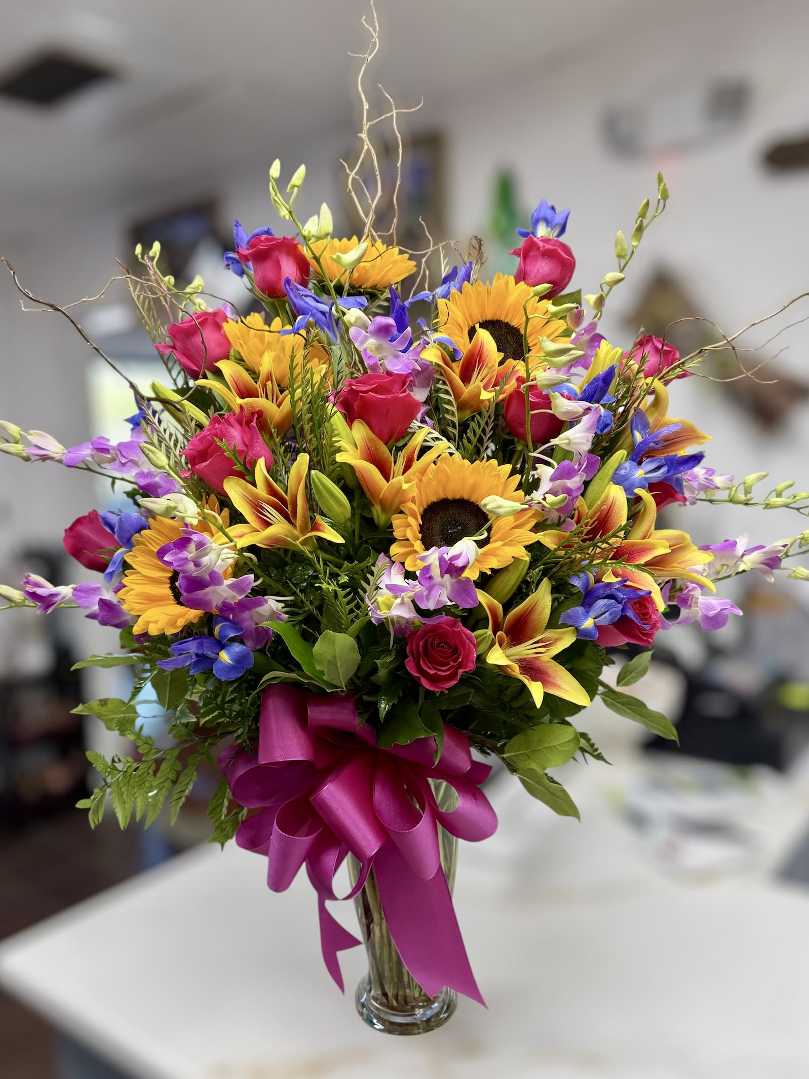 Tall mixed bouquet with sunflowers, pink roses, lilies and purple-blue blooms in a glass vase with magenta bow