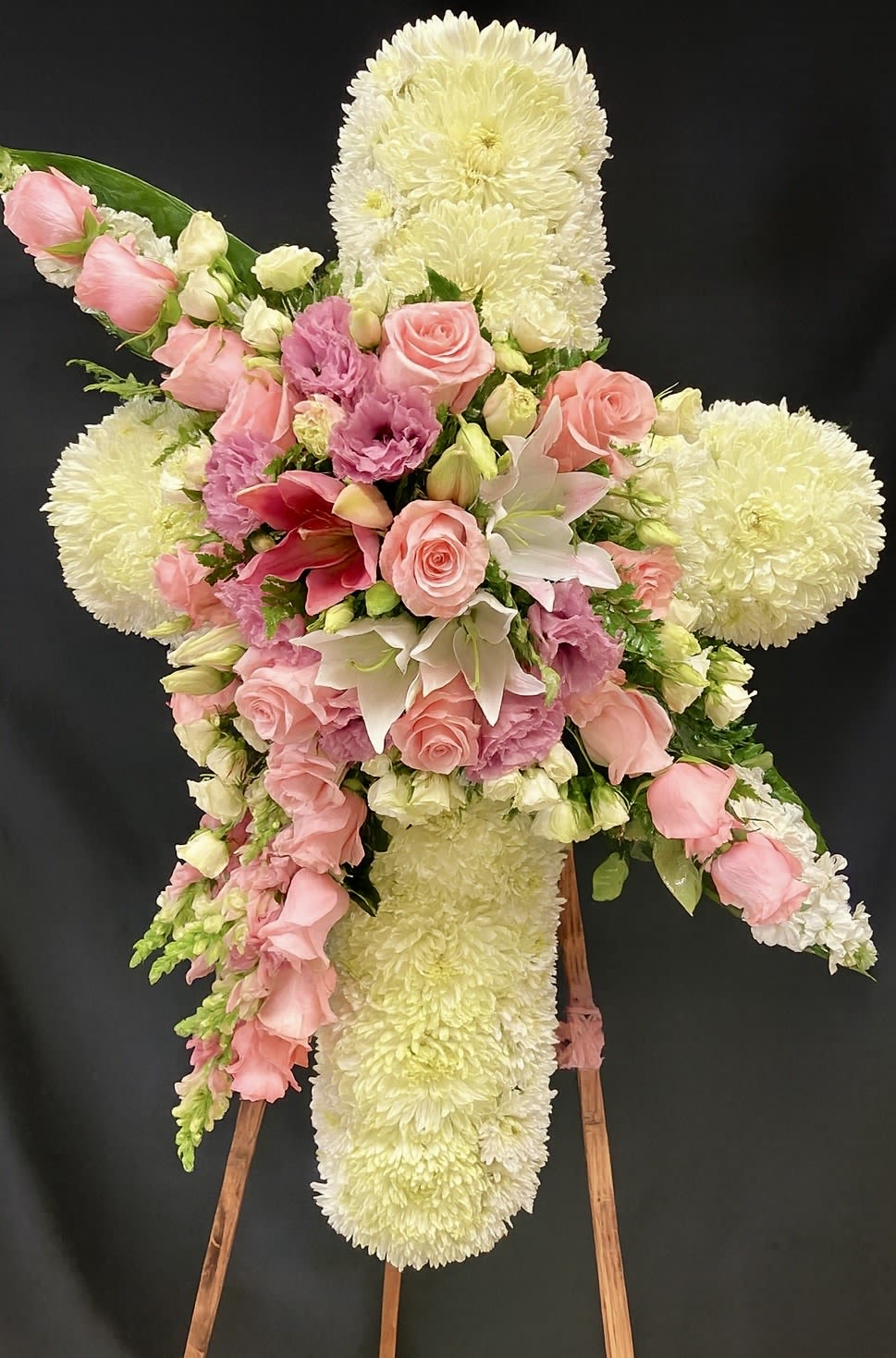 Standing floral cross of white mums with cascading pink roses, lilies, and lavender blooms on a wooden easel.
