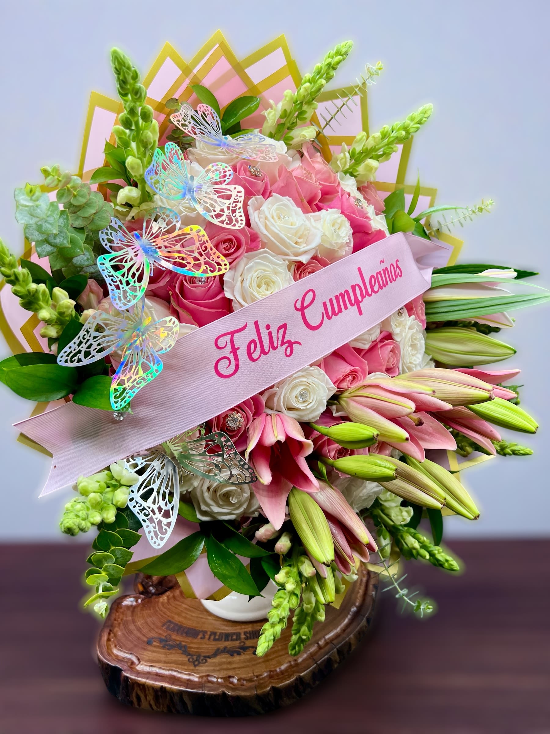 Tall arrangement of pink lilies and pink and white roses with a Feliz Cumpleaños ribbon and butterfly accents.