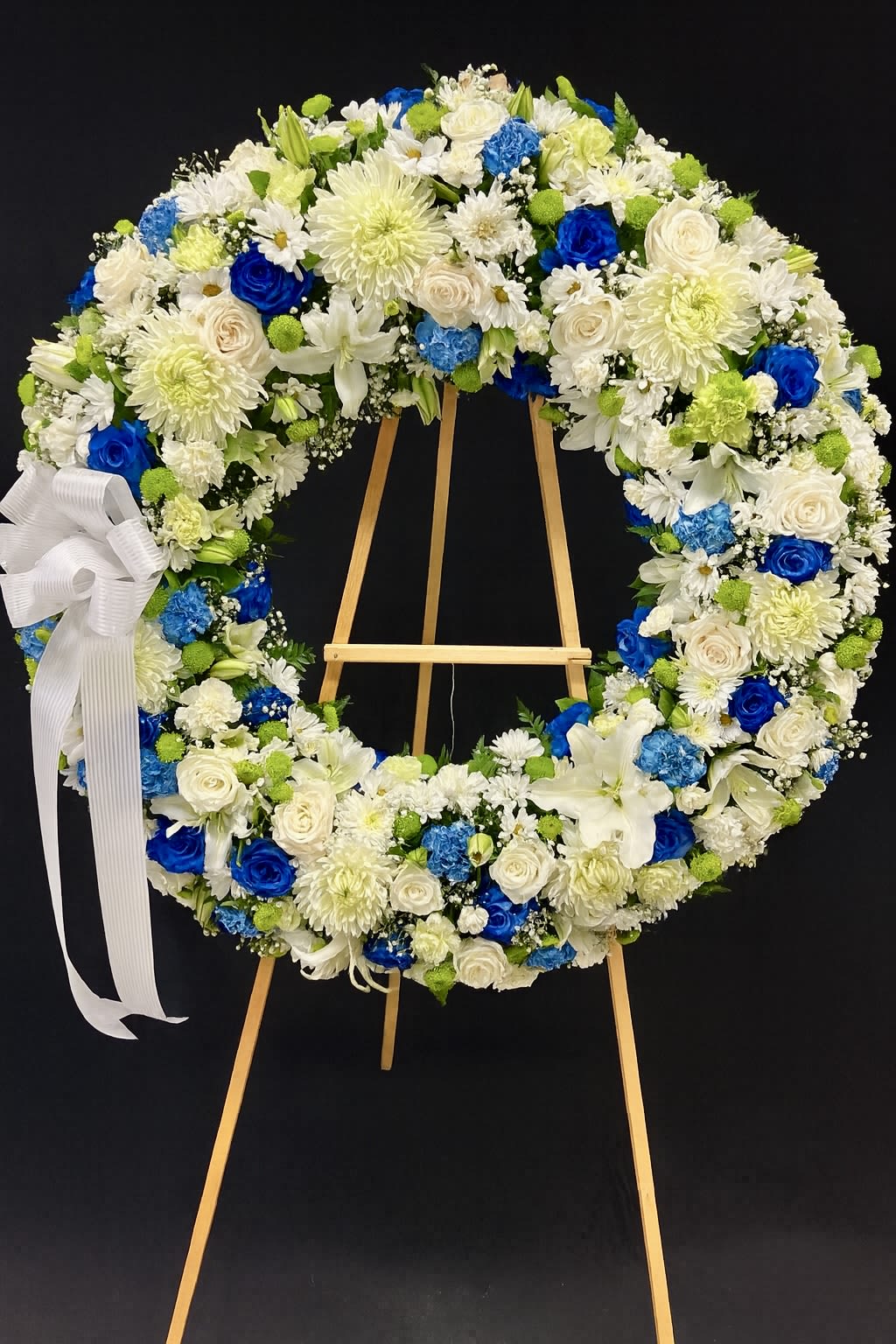 Large standing wreath of white and blue flowers with a white ribbon bow on a wooden easel