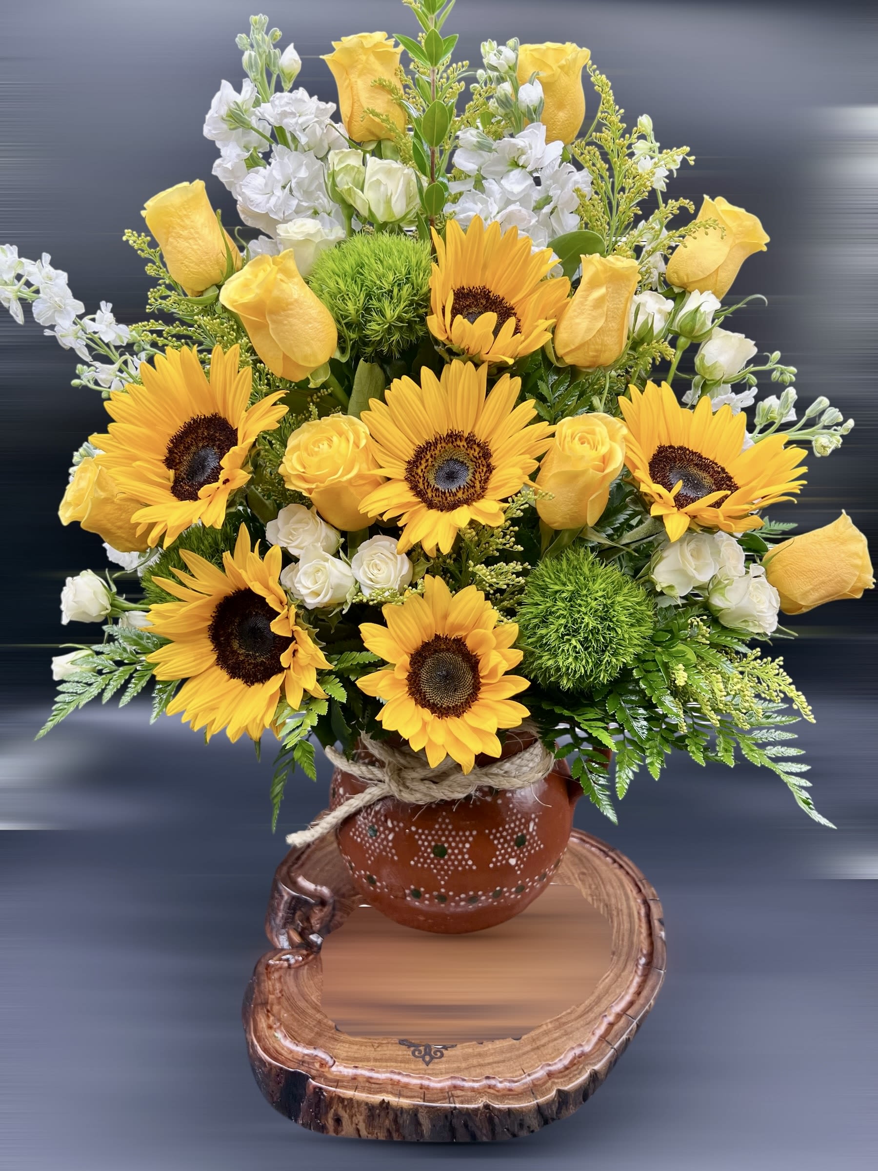 Arrangement of yellow sunflowers and roses with white blooms in a rustic clay pot on a wood slab base