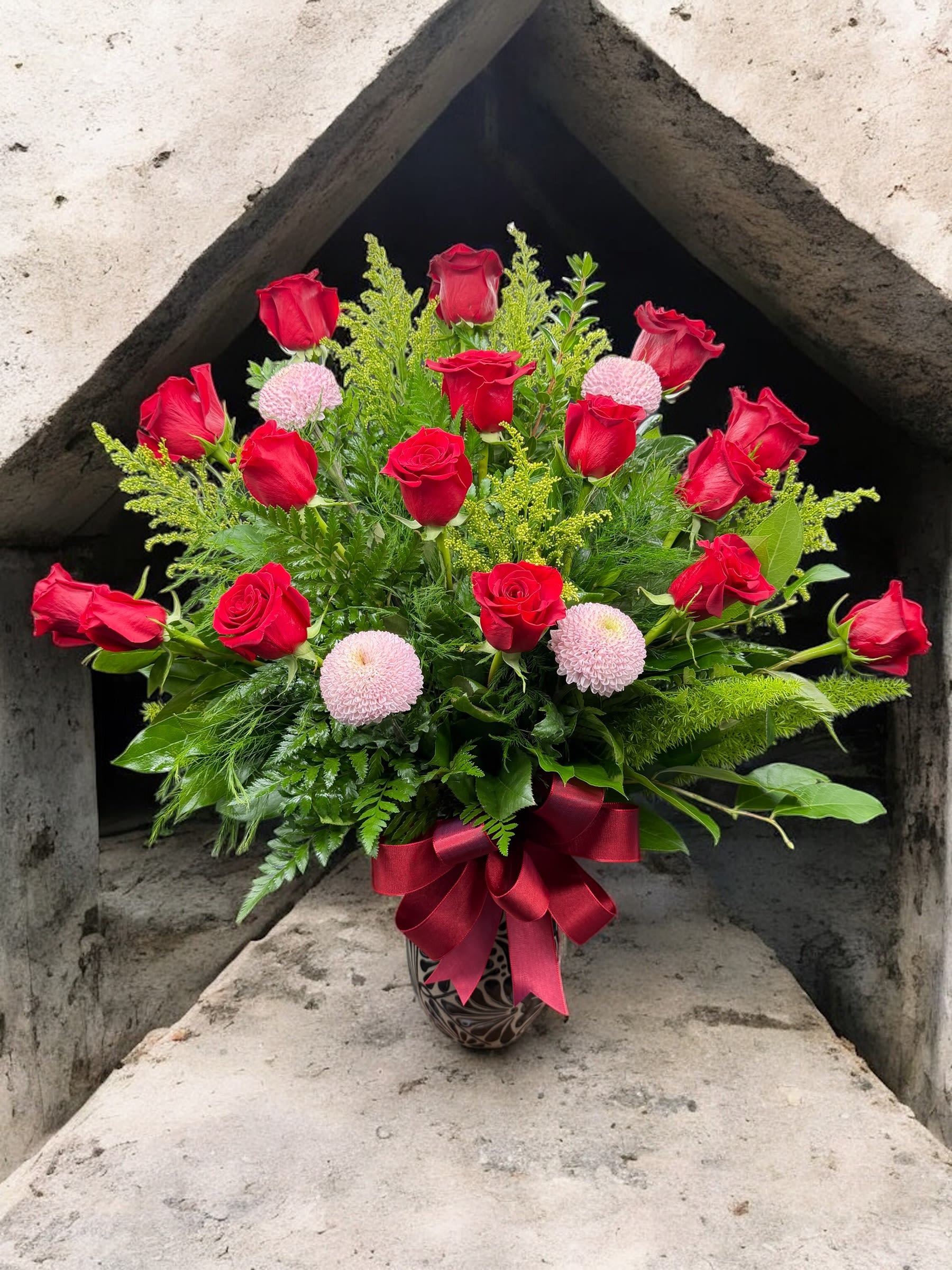 Red roses and pink pompom flowers in a patterned vase with a burgundy ribbon