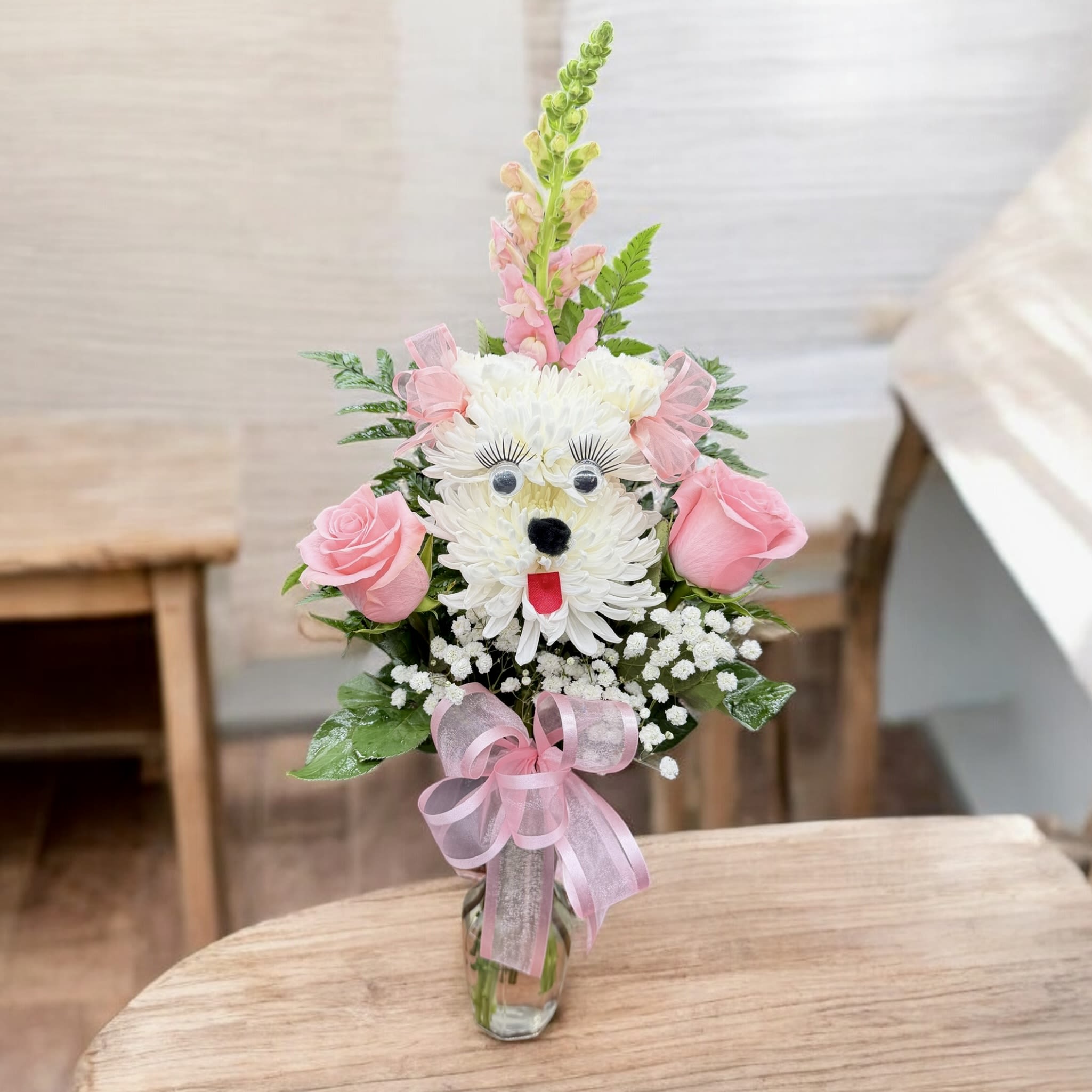 Doggy’s Face - A cute and playful floral “pup” arrangement designed with white mums to form the face, complete with adorable eyes and lashes, a little nose, and tongue. Accented with soft pink roses, baby’s breath, fresh greenery, and a tall snapdragon for height, then finished with sweet pink bows and a pink ribbon on a clear vase—perfect for birthdays, get-well wishes, or just to make someone smile.  COLOR CAN BE CHANGE AS CUSTOMER REQUEST.