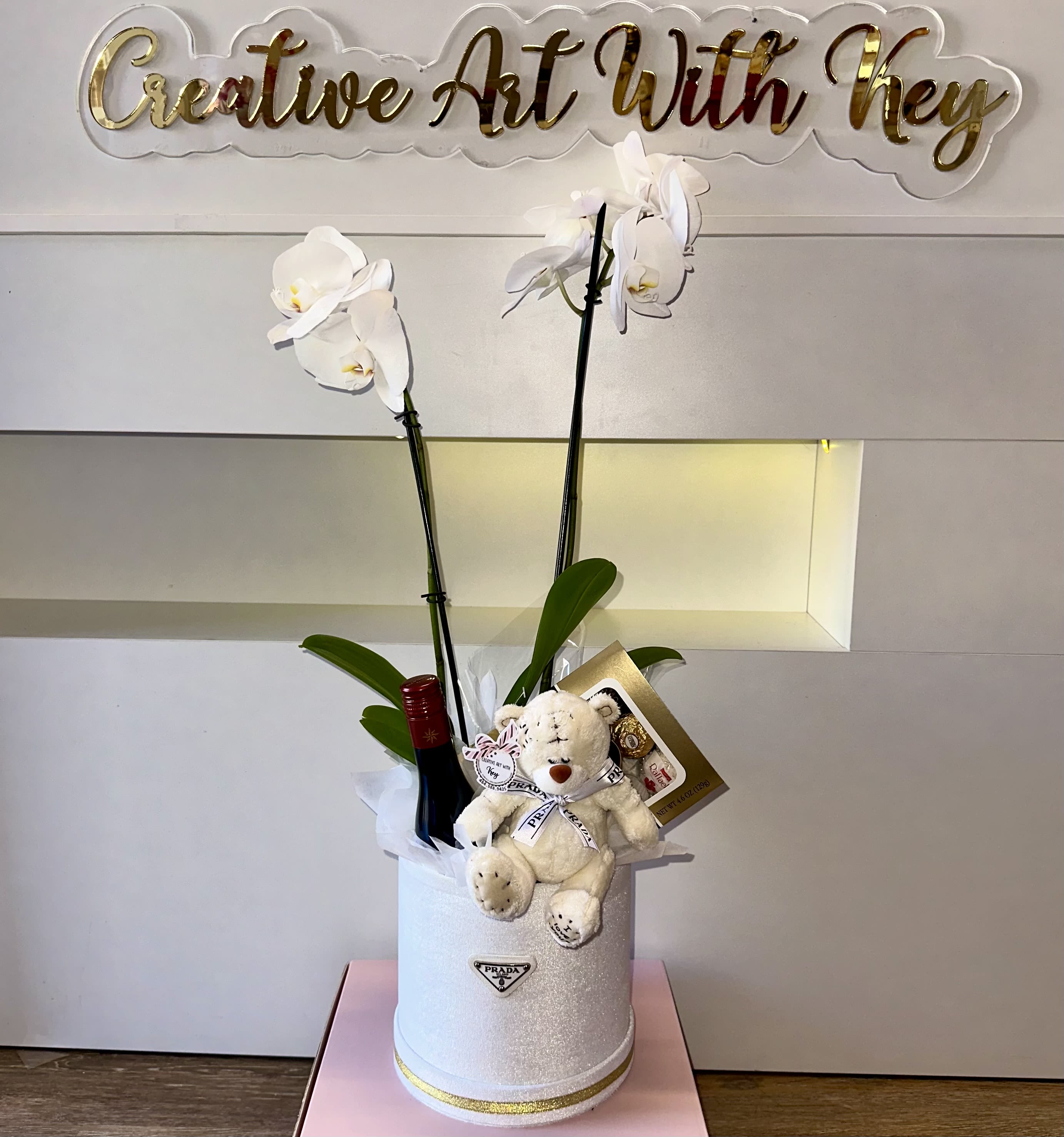 White orchids in a glittery box with a teddy bear and wine bottle