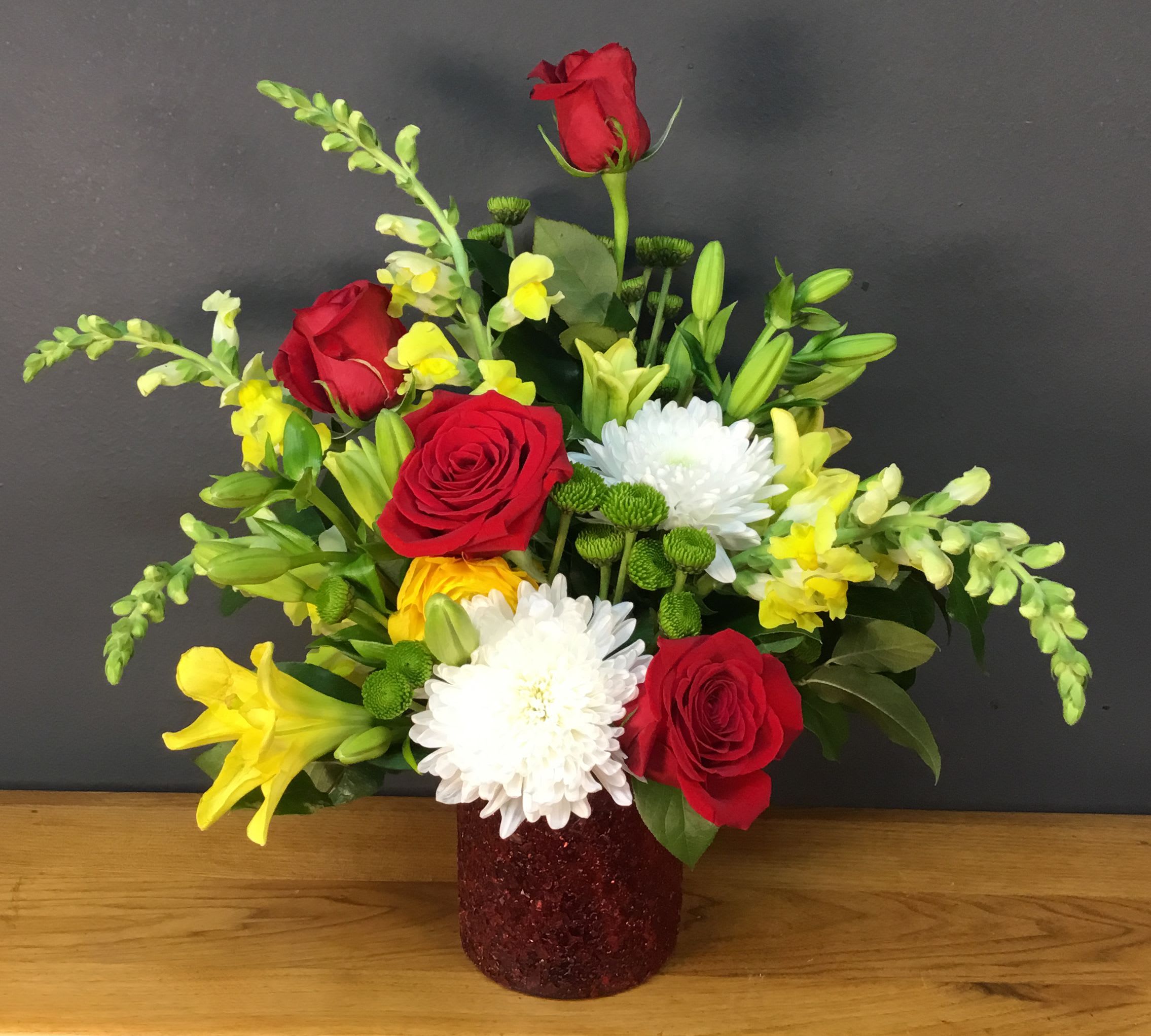 Yellow Brick Road - Bright, celebratory, and full of texture - this Yellow Brick Road flower arrangement is a cheerful bouquet perfect for a birthday, thank-you, or just-because surprise. The arrangement pairs red roses and white chrysanthemums with sunny yellow lilies and pale yellow snapdragon spikes so the person you're thinking of will feel genuinely celebrated the moment it arrives.  Flowers are arranged densely with bright green accents for lively contrast, creating a compact, layered look that reads joyful from every angle. Everything sits in a red glitter vase that gives the display a festive, jewel-toned base and a slightly formal sparkle - ideal for tabletops or office desks. The mix of velvety rose petals, ruffled chrysanthemum blooms, the trumpet shape of lilies, and vertical snapdragon spikes provides both visual variety and pleasant texture.  Perfect for everyday gifting or marking a special moment, this design sends a warm, upbeat message without being over the top. Because it's compact and staged for immediate impact, it's easy to place and enjoy right away; standard floral care - fresh water and a cool spot out of direct sun - will keep the blooms looking their best.