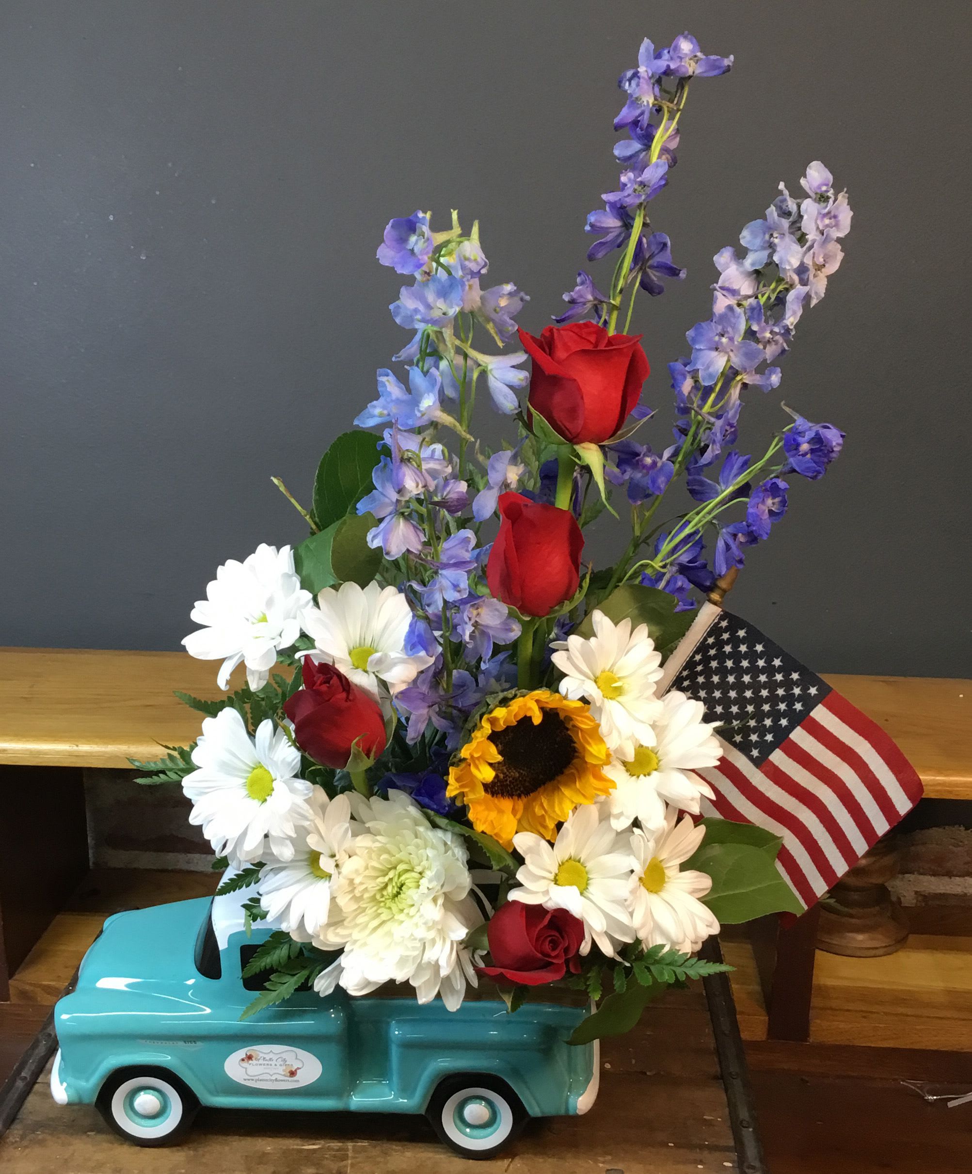Proud to be American - A cheerful, Americana-inspired flower arrangement that says "celebrate" with a wink - perfect as a birthday bouquet, a Father's Day pick-me-up, or a bright "just because" surprise. This floral arrangement places red roses, white daisies and chrysanthemums, and blue‑purple delphinium into a whimsical turquoise ceramic truck container, finished with a small American flag for a festive touch that also works for Fourth of July or congrats moments. The mix reads patriotic without feeling formal, so the person you're thinking of will feel both honored and delighted.  