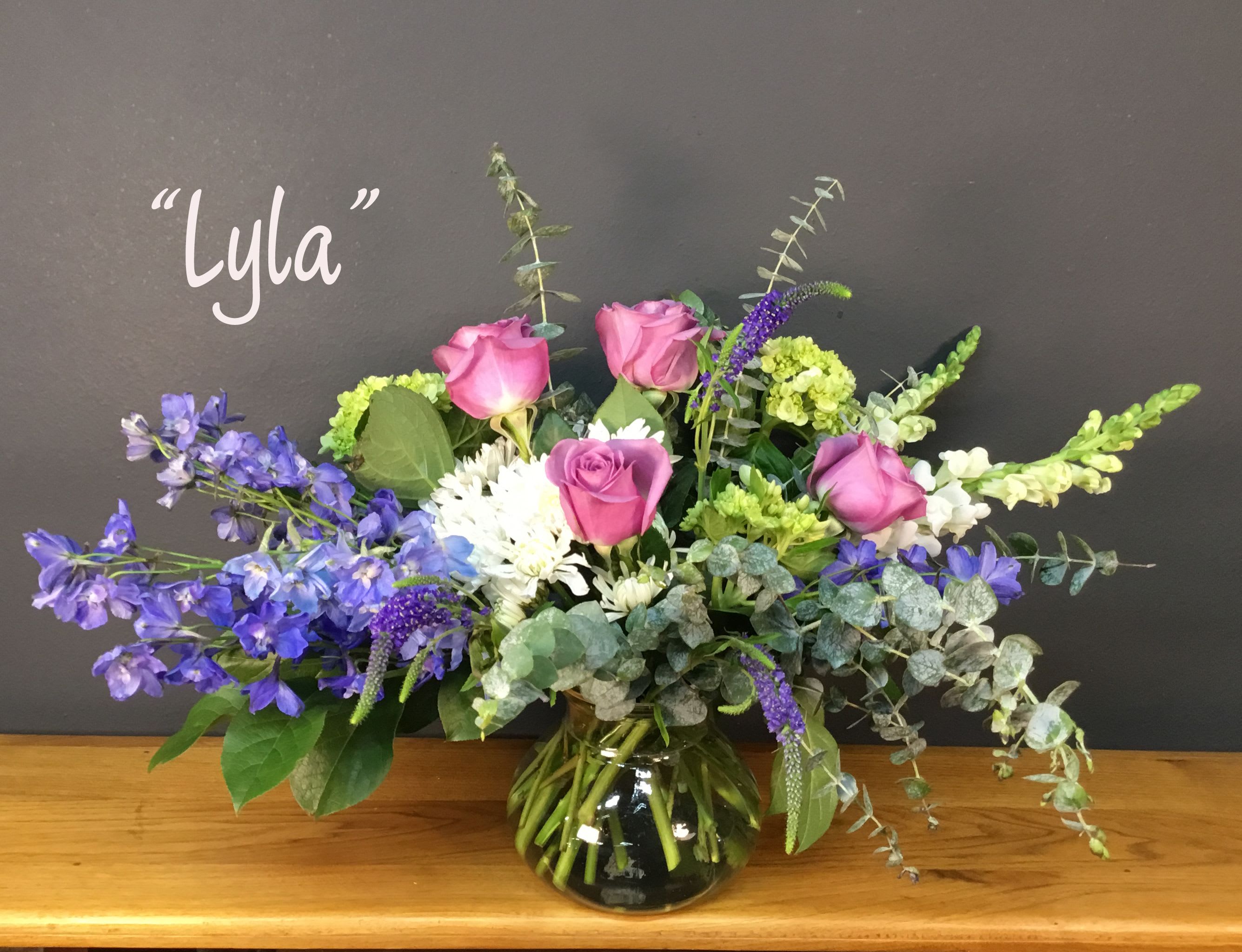 "Lila" designed by Marla Storey - Pink roses paired with blue-purple delphinium and tall snapdragon spikes give this flower arrangement a fresh, garden-fresh personality that delights for a birthday, a thank-you, or a spring celebration. The mix of pink, purple, white and green creates a cheerful, uplifting look that reads both elegant and playful-the kind of bouquet that makes the person you're thinking of feel genuinely celebrated.  