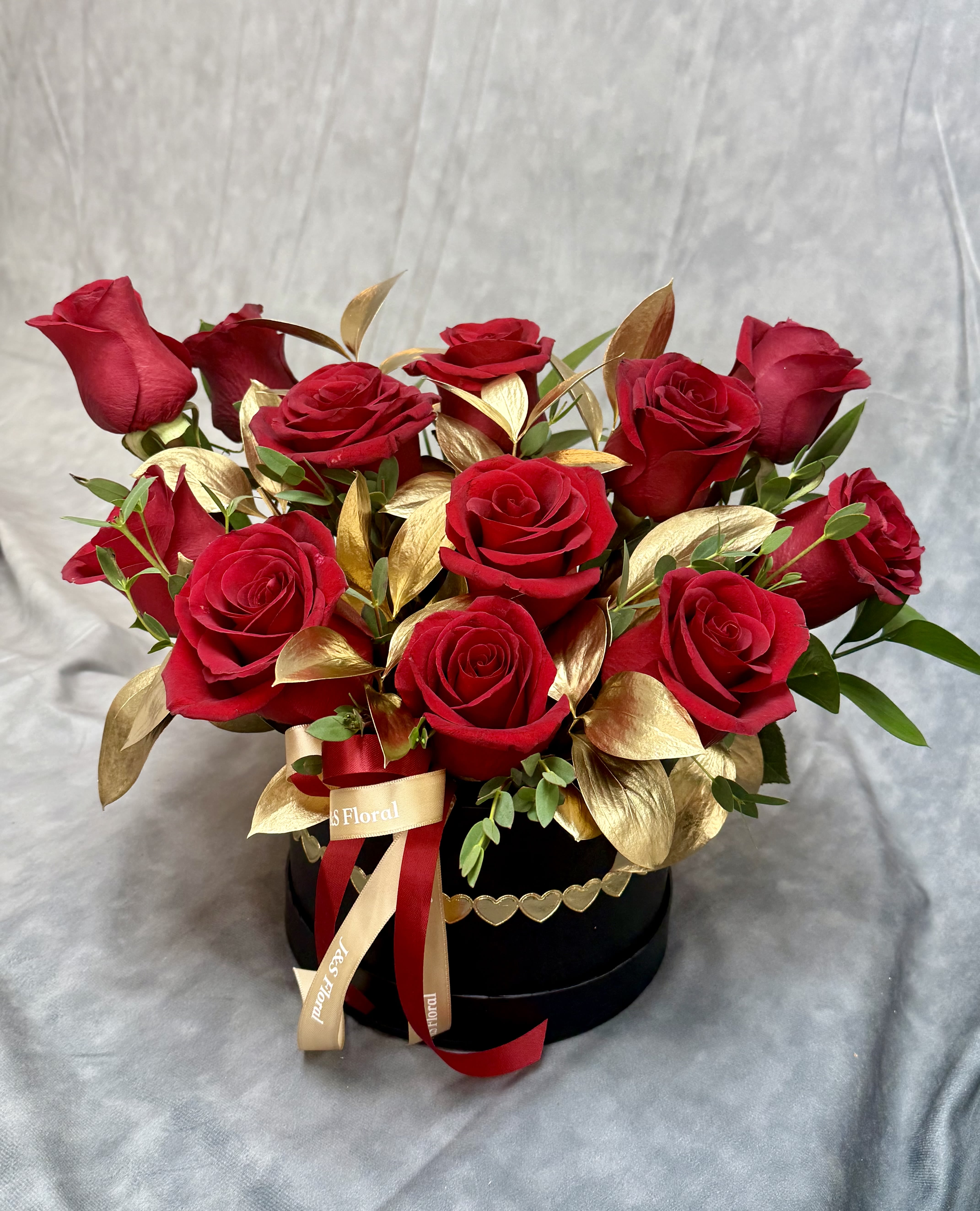 Red roses arranged in a black round box with gold leaves and ribbon