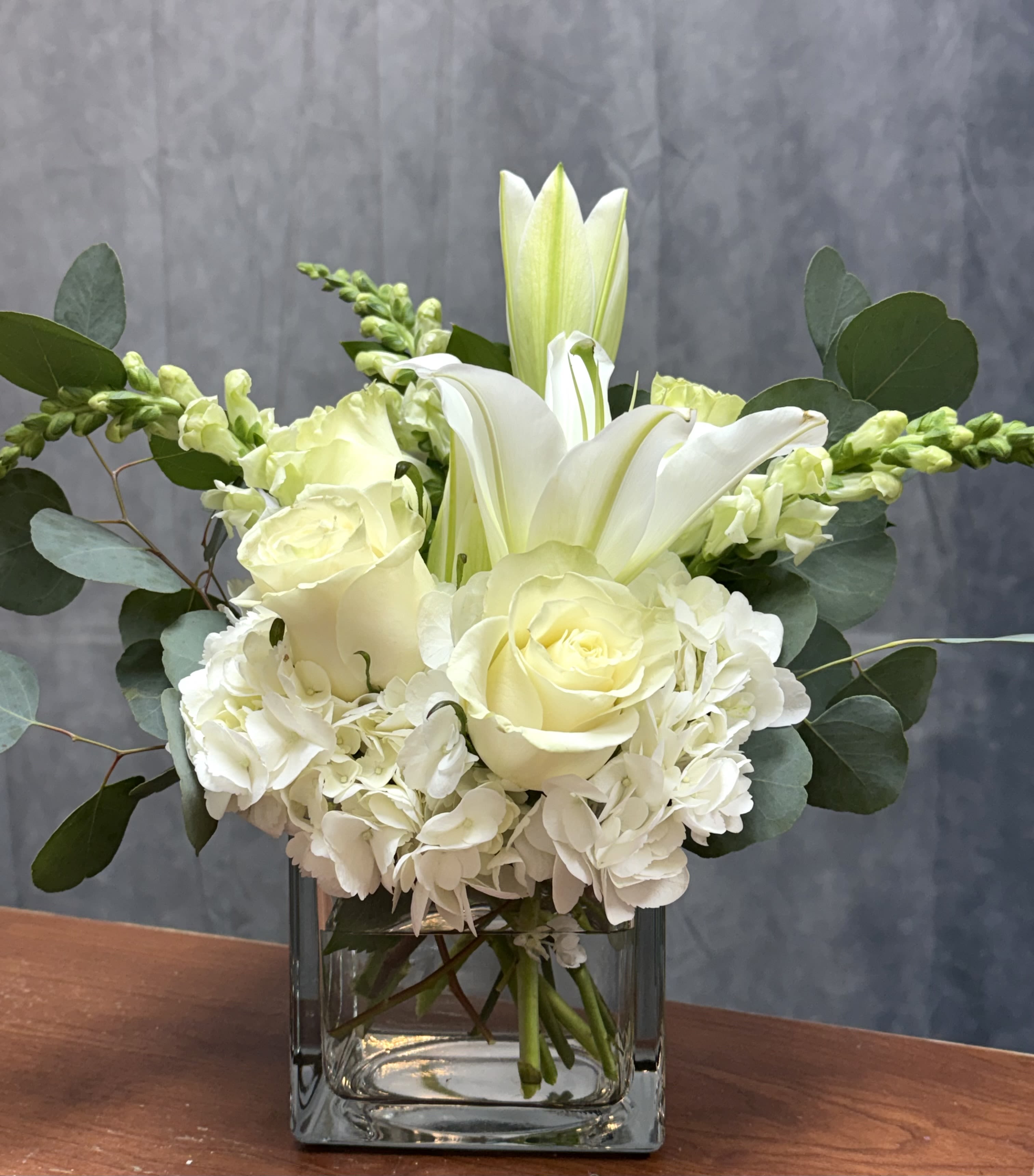 White lilies, roses, and hydrangeas arranged in a clear glass cube vase