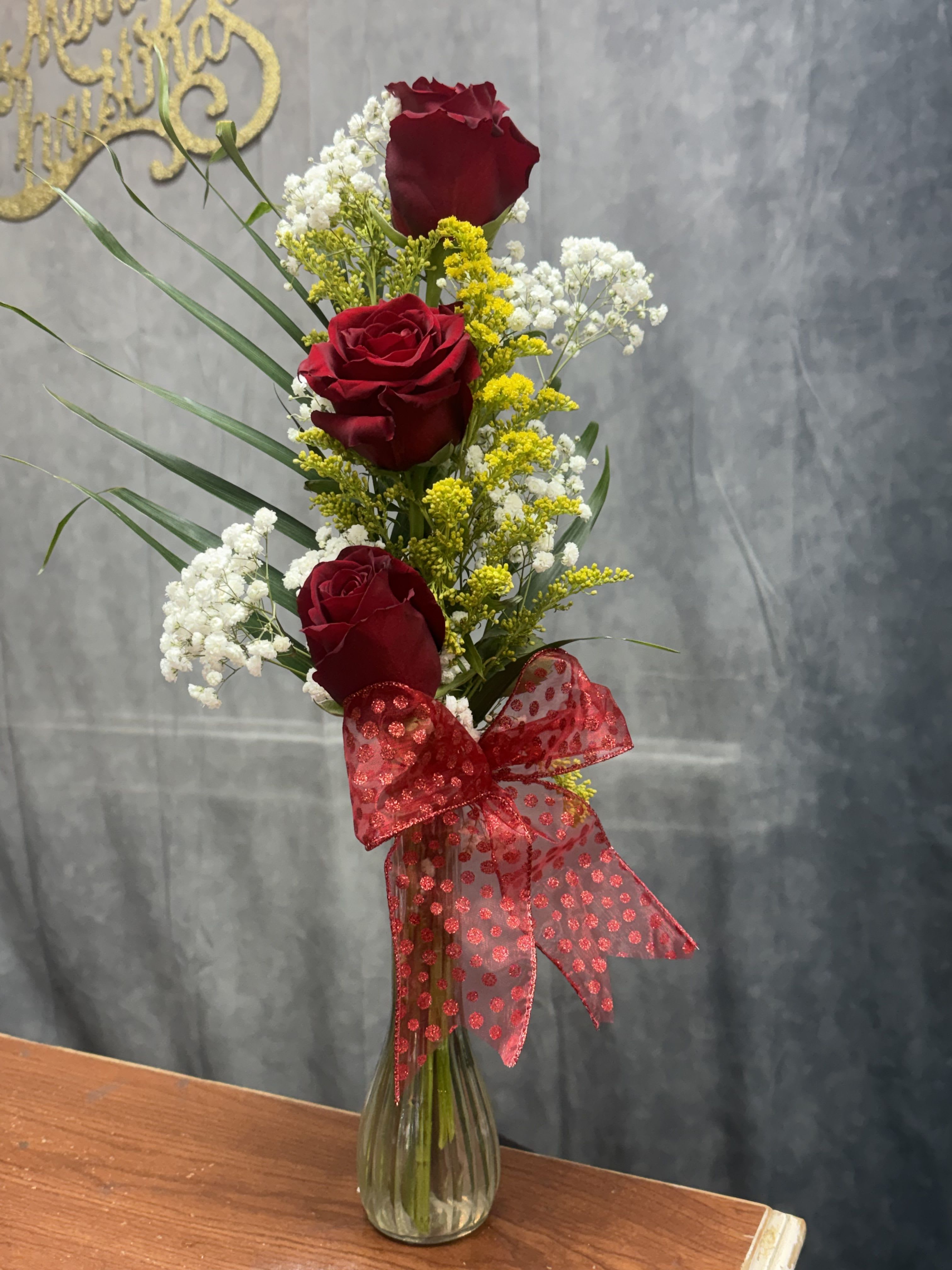 Three red roses in a glass vase with white and yellow filler flowers and a red ribbon