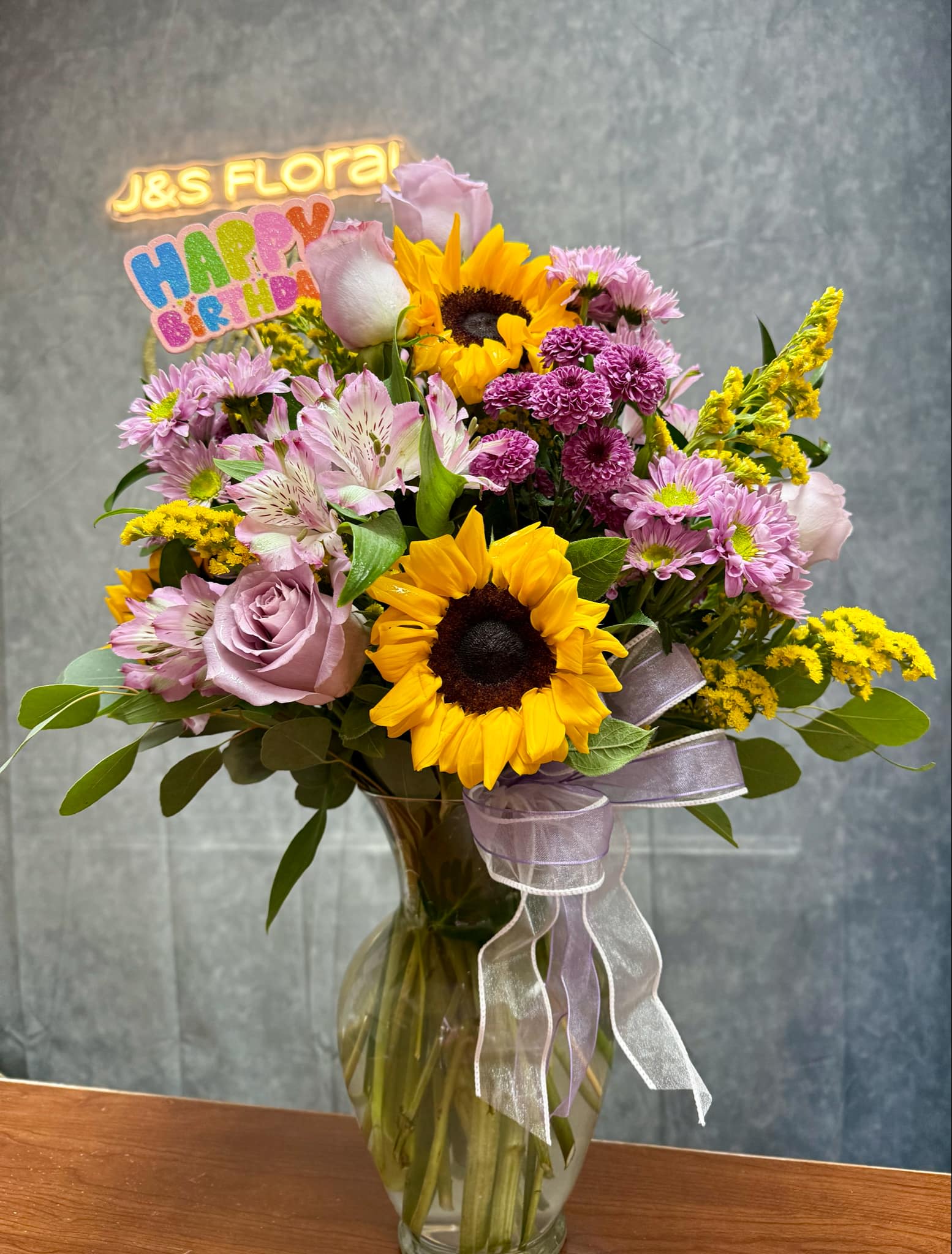 Mixed bouquet with sunflowers, pink roses, and purple blooms in a glass vase