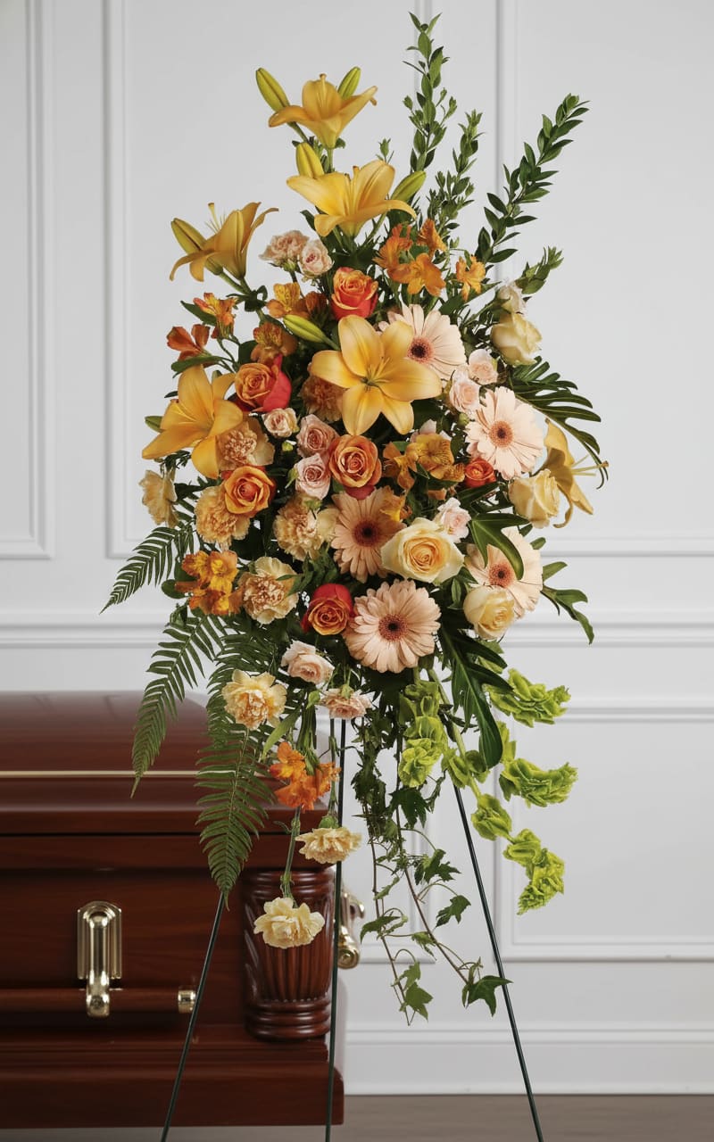Tall standing spray with yellow lilies and peach roses on an easel beside a wooden casket.