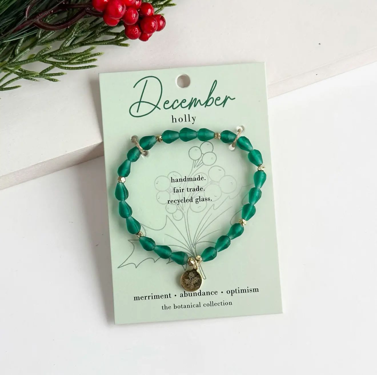 December Birth Flower Bracelet  - Hello, December!  Part of our Botanical Collection, the December Bracelet is crafted from rich green glass beads, brass accent beads, and features a holly charm. Great for gifting, the card features the holly and it's symbols of merriment, abundance and optimism, along with a garden-inspired quote.This bracelet also benefits an urban community garden project!  The semi-opaque beads have a satin, frosted texture and are handmade from recycled glass. The flower charm is 24k gold plated.  Sustainably handmade by women artisans in India.  