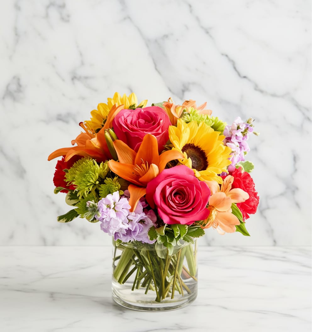 Floral Embrace - Bright, cheerful color bursts make this mixed flower arrangement a joyful pick for a birthday bouquet, thank-you gift, or simply to let someone know you're thinking of them. The rounded, compact design places hot pink roses, sunny yellow sunflowers, and vibrant orange lilies front and center, while pale lavender stock and soft purple fillers add cool contrast that keeps the palette lively rather than overpowering.  Carefully packed for a lush, abundant look, the arrangement also includes red and pink carnations, chrysanthemums and other seasonal blooms layered together for texture and fullness. It arrives in a clear glass vase that highlights the stems and keeps the composition feeling fresh and garden-like; the size and dense styling make it an eye-catching centerpiece or a single-surface statement that will brighten a kitchen table or office desk.  Perfect for birthdays, congratulations, or an everyday "you matter" surprise, this bouquet is designed to make the recipient feel celebrated and seen. Standard vase care-fresh water and a cool spot away from direct sun-will help the mixed stems stay vibrant for days.