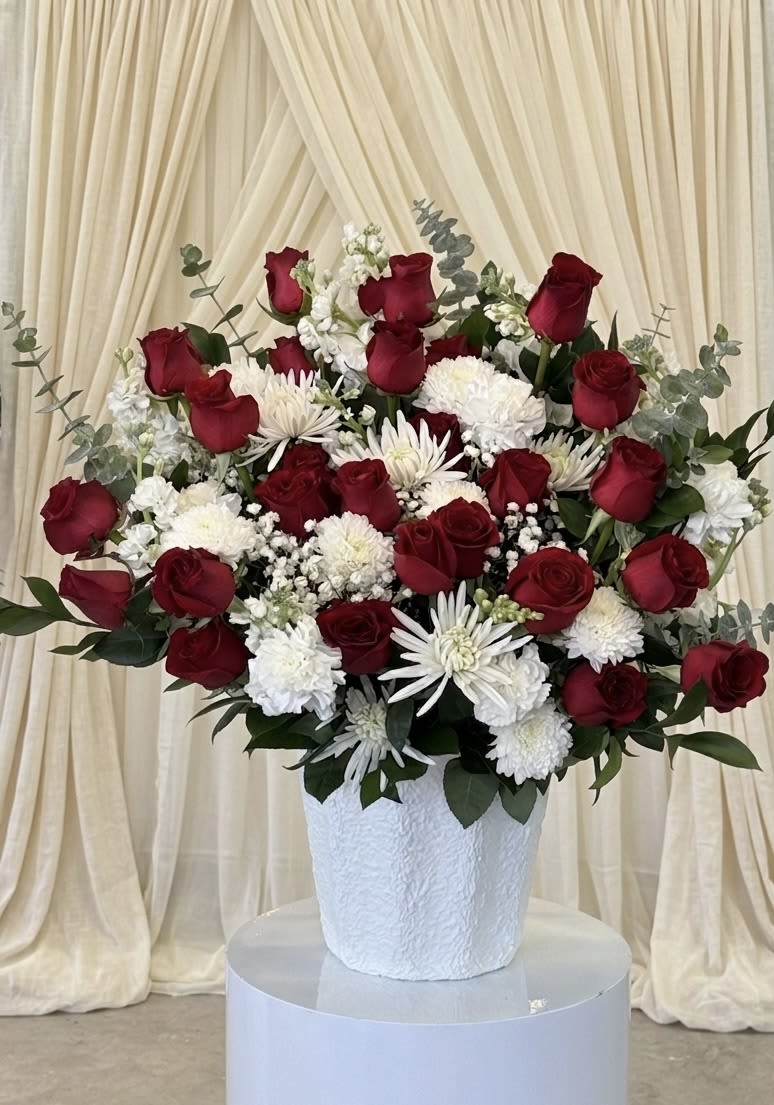 Endless Love. By Anita - Dense clusters of deep red roses paired with crisp white blooms create a romantic, ceremonial centerpiece perfect as an anniversary roses arrangement or a sympathy tribute. The mix of velvety red roses and round white chrysanthemums, accented by spiky white flowers and gray‑green eucalyptus-like foliage, gives the person you're honoring a feeling of timeless devotion and quiet elegance.  Arranged symmetrically in a white textured vase on a round pedestal stand with cream drapery behind, the composition reads formal and polished-ideal for weddings, memorial services, or any moment when you want to express enduring love. The full, compact shape and contrasting red, white, and green palette make it easy to place as a focal centerpiece or tribute; minimal care is needed beyond fresh water in the vase to keep the blooms looking their best.