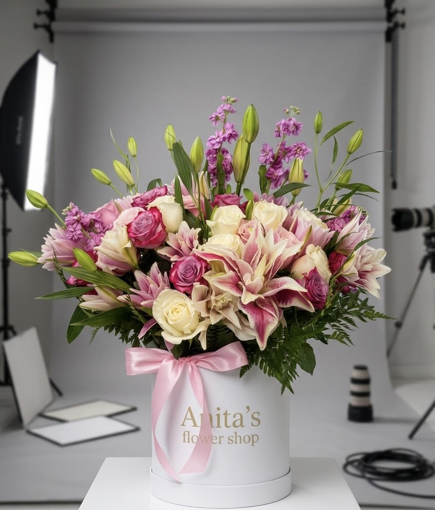 Pink  Velvet Box. - Soft pink and cream lilies rising above a cloud of roses and tiny purple blooms create a romantic birthday or anniversary flower arrangement that feels both luxurious and intimate. This flower arrangement for Mother's Day or a special celebration is presented in a white cylindrical hatbox, the pink satin ribbon tied around it giving the gift a polished, ready-to-present look that will make the person you're thinking of feel truly celebrated.  Densely layered for a full, rounded silhouette, the design mixes pink and white lilies, matching roses, and small purple filler flowers with touches of green for contrast and texture. Several upright lily buds add a slightly dramatic height while keeping the overall profile lush and compact - perfect for tabletops or a bedside table. The hatbox keeps stems neatly contained and arrives gift-ready with the satin ribbon; water and care instructions included by request will help the recipient enjoy the blooms longer.
