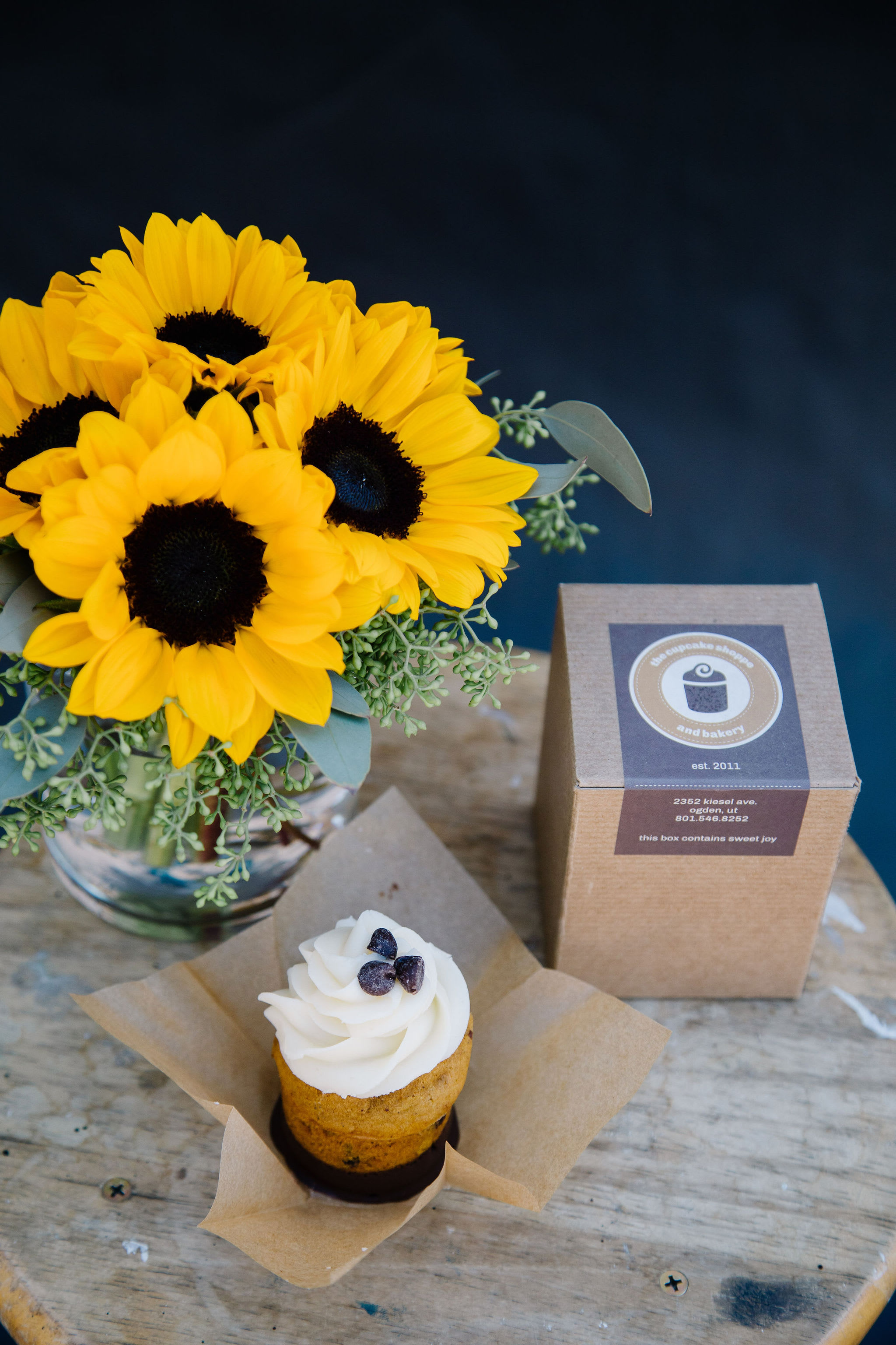 A Sweet Fall - 6 mini sunflowers with eucalyptus accents hand-tied in a short cylinder vase paired with a single large artisan cupcake from the Cupcake Shoppe &amp; Bakery; flavors vary daily.