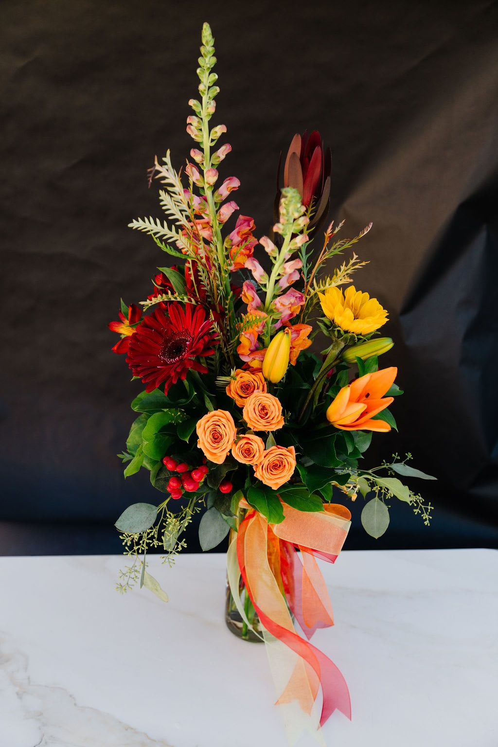 Autumn Embrace - Warm and elegant, Autumn Embrace combines the beauty of deep red gerbera daisies, golden sunflowers, vibrant orange roses, lilies, and snapdragons accented with lush greenery and berries. Finished with a flowing ribbon in autumn hues, this arrangement is perfect for celebrating the season, brightening a table, or sending a heartfelt gift.