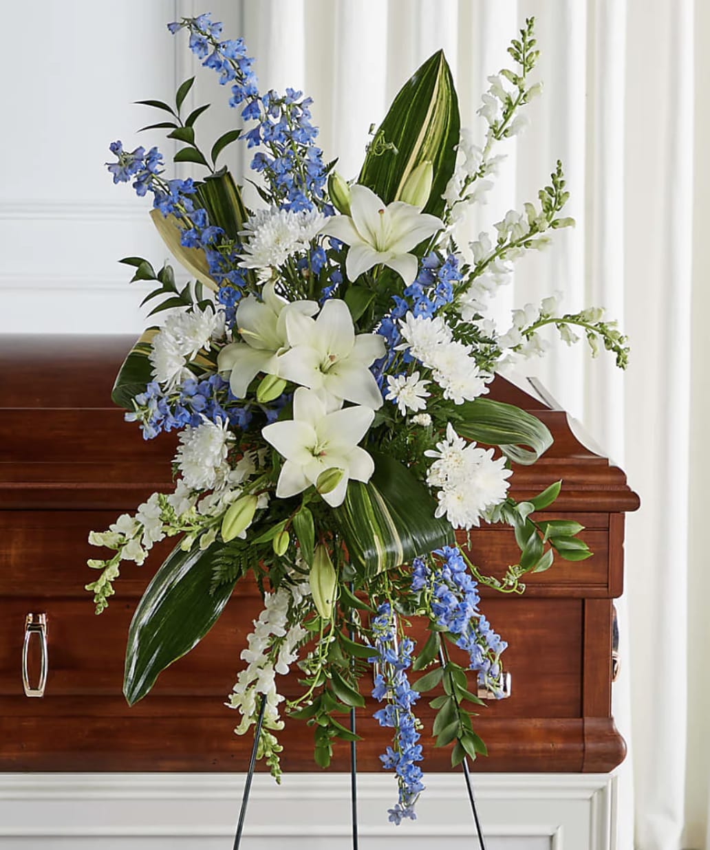 Heaven's Promise Standing Spray - Honor a cherished life with the timeless elegance of Heaven's Promise Standing Spray. Featuring pristine white lilies, vibrant blue delphinium, graceful white snapdragons, and premium seasonal blooms, this striking tribute symbolizes peace, faith, and enduring love. Handcrafted by the floral designers at Bloom Luxury Box, each arrangement is thoughtfully designed to offer comfort and support while celebrating a life beautifully lived. Displayed on an easel, this elegant standing spray provides a meaningful expression of sympathy and a lasting tribute to treasured memories.