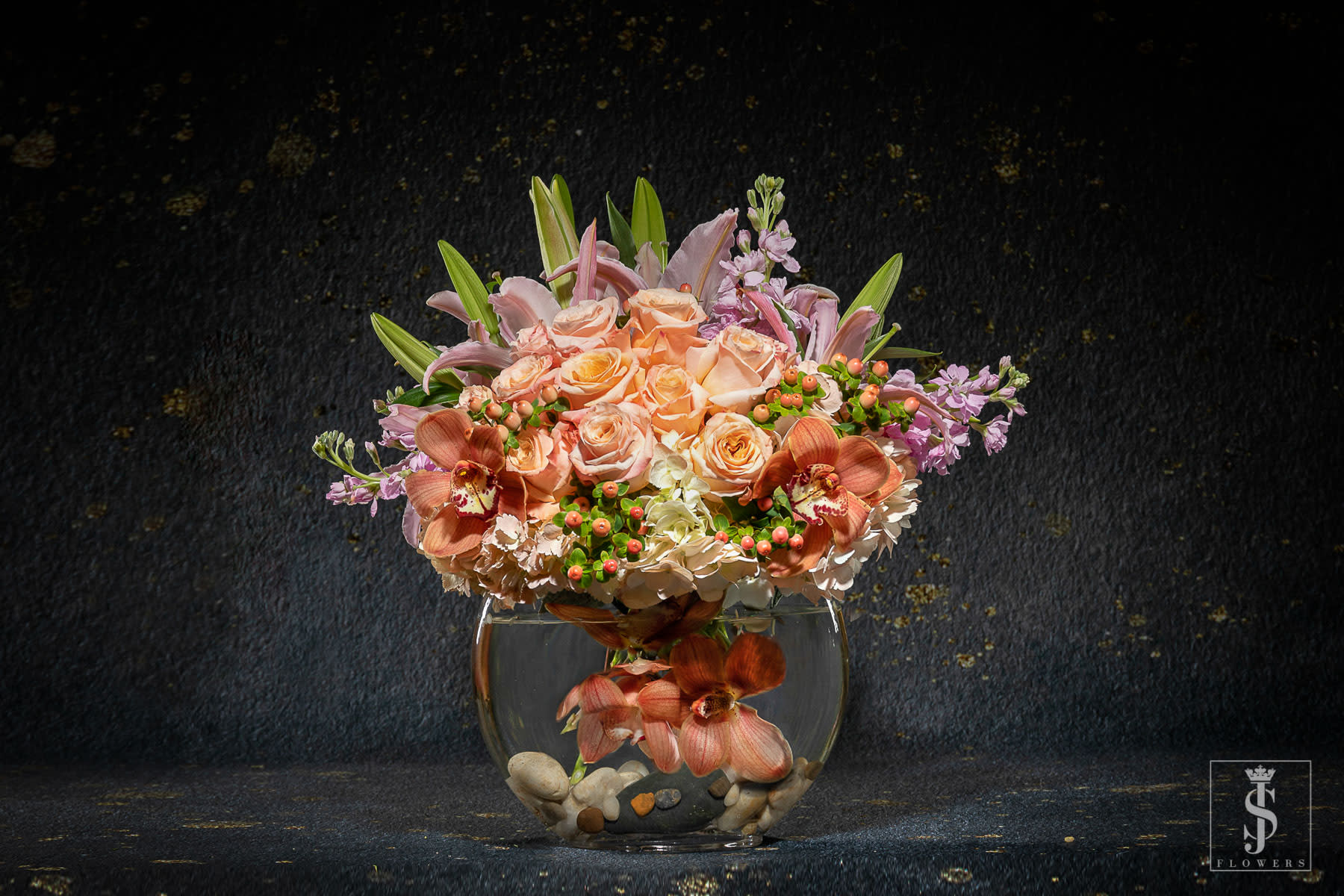 Peach roses and orchids arranged in a round glass vase with stones