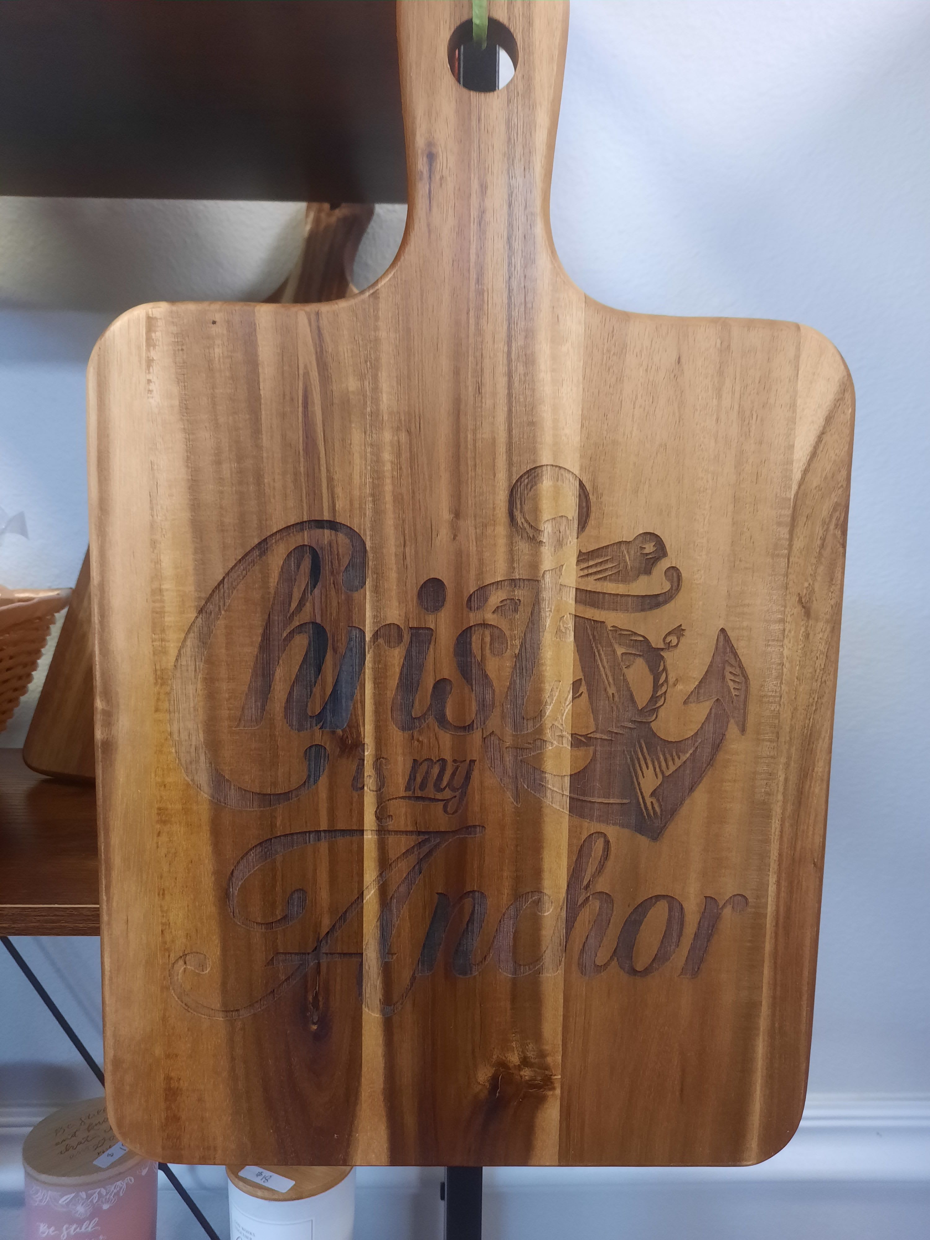 "Christ is my Anchor"  Joe McShan Cutting Board  - A rustic wooden paddle board engraved with "Christ is my Anchor" creates a warm, faith-centered accent for home or gifting moments. As a thoughtful Christian gift or church decoration, this piece reads clearly as a message of steady faith-perfect for baptisms, new-home blessings, or to encourage someone facing change.  The paddle is rectangular with a convenient handle and hanging hole at the top for easy display. Natural warm-brown wood grain shows through a contrasting dark engraving of the phrase and a simple anchor illustration,  and readable. Its size and flat profile make it easy to tuck onto a gallery wall, lean on a shelf, or hang in an entryway.  Give this paddle to the person you want to remind that their faith holds true-an understated, comforting statement piece that fits cottage, coastal, and farmhouse decor. Hang it straight from the included hole; no special care is required beyond keeping it dry and dust-free to preserve the wood finish.