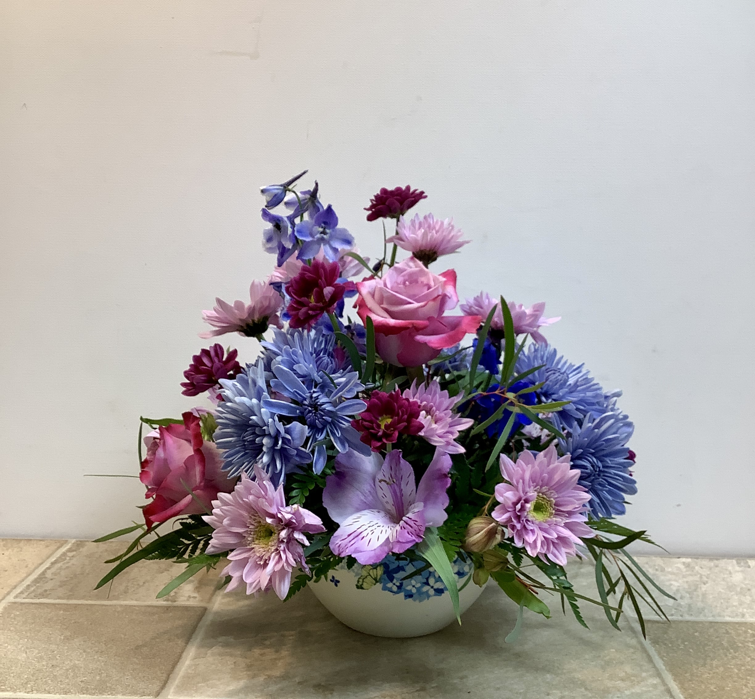 Periwinkle Promise - This garden-style floral arrangement offers a cohesive color palette of blues, purples, and pinks, arranged in a decorative bowl that complements the floral colors.  Purple roses are surrounded by charming blue chrysanthemums, light lavender-pink alstroemeria, and purple chrysanthemums. Stems of blue delphinium add vertical interest, and wispy, fine greenery is tucked throughout, giving the arrangement an organic, "just-picked" feel.  Approximate size: 13" w x 13" h  Primary flowers: rose, mum, lily