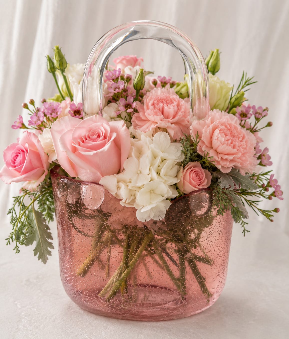 Petal Perfection's "Let my get my Purse" in Pink - A playful cascade of soft pink roses and coral carnations spills from a clear pink, handbag-shaped glass vase-this flower arrangement is a joyful pick for a birthday bouquet, anniversary surprise, or a just-because gift that reads like a warm, affectionate hug. The pastel mix of pink, red, peach, white, purple, and glossy green foliage creates a romantic, cheerful mood perfect for spring, Mother's Day, or any moment when you want someone to feel truly seen.  Dense, rounded clusters of roses, carnations, and small daisy-like blooms are balanced by upright snapdragon spikes and the lacy texture of white hydrangea for height and movement; small pink filler flowers tuck into the mix for a finished, feminine silhouette. The handbag glass vase makes the arrangement instantly gift-ready and playful-flowers rise above the handle for a whimsical, overflowing look that delights at first glance.  Made to celebrate moms, partners, friends, or coworkers, this arrangement sends an unmistakable message of affection and thanks. To keep it fresh, change the water regularly and trim stems every few days; choose your vase option at checkout if you prefer an alternative presentation for immediate gifting.