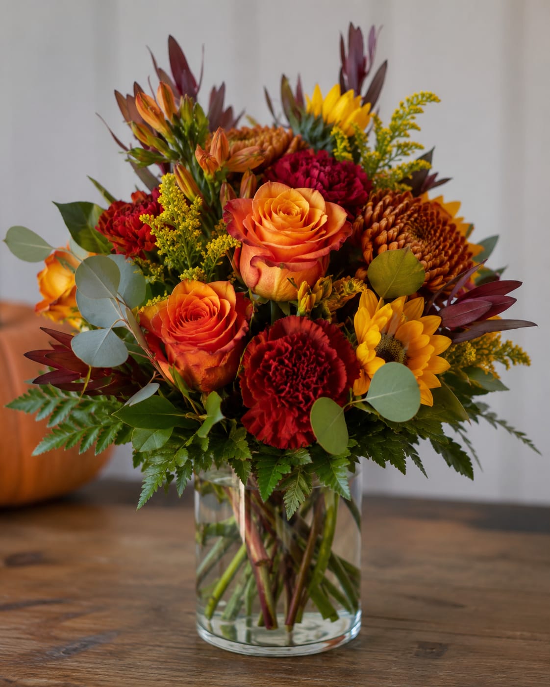 Possie Bliss  - A rich, autumnal display of color greets the eye-this flower arrangement brings together orange roses, deep red carnations, sunny yellow sunflowers, and warm chrysanthemums in a clear glass vase for a fall birthday bouquet or a heartfelt thank-you gift. The palette moves from bright golden yellow through burnt orange to burgundy, creating a cozy, celebratory vibe that makes the person you're thinking of feel warm and seen.  The mix is densely arranged for a full, abundant silhouette: rounded roses and chrysanthemums are offset by spiky alstroemeria accents and airy filler for layered texture and strong contrast. Presented in a simple clear cylindrical glass vase, the proportions keep the focus on the blooms while letting stems show through for a clean, modern touch. Ideal for autumn gifting, seasonal décor, or to brighten someone's day with vivid, comforting hues.