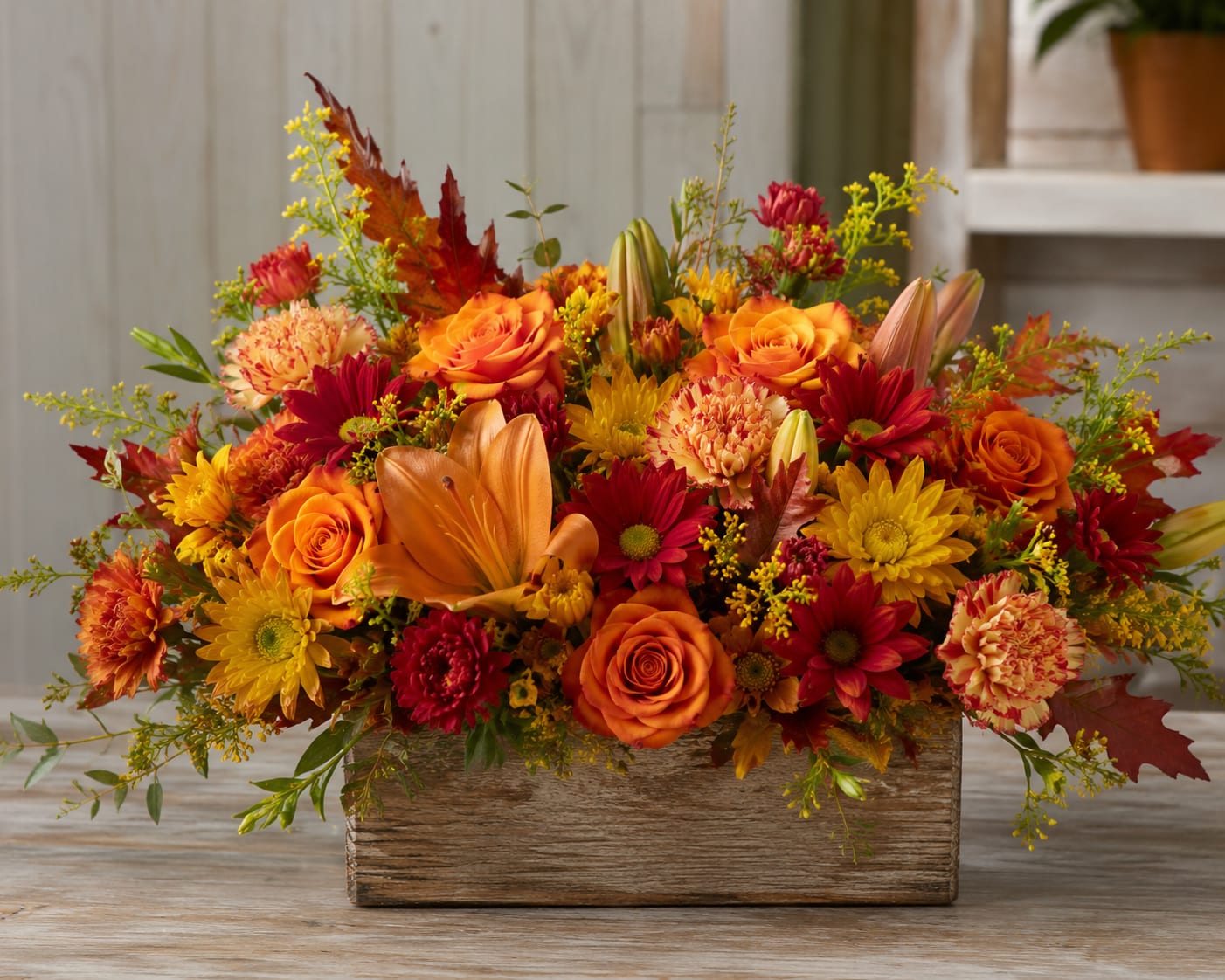 Rustic Fall for Tampa  - A dense, low centerpiece that bursts with warm autumn color-this flower arrangement for fall and Thanksgiving brings the cozy, handcrafted charm of a Rustic Fall for Tampa to any table or entry. Perfect as a Thanksgiving centerpiece, seasonal gift, or birthday surprise for someone who loves autumn hues, the design reads cheerful and heartfelt so the person you're thinking of will feel celebrated and seen.  Packed into a wooden box for a rustic, farmhouse feel, the arrangement layers orange roses, peach-toned carnations, red daisy-like blooms, yellow chrysanthemums and cheerful daisies with a few lily buds and striking broad orange lily petals rising above the mix. Small yellow filler sprays and scattered fall leaves add texture and movement, creating a compact, garden-fresh look with rich orange, red, yellow and pink accents.  Ideal for the host who wants a low, conversation-friendly centerpiece or for brightening a bedside or kitchen table, this sturdy wooden-box arrangement requires minimal care-keep in fresh water, trim stems if needed, and place out of direct midday sun to extend vase life. Choose this when you want a warm, seasonal gift that reads like a thoughtful harvest hug.