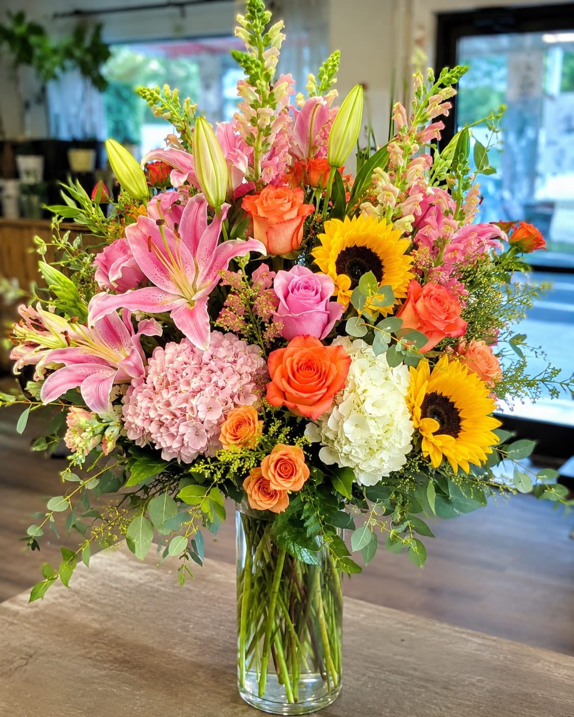 Tampa Bay Bliss by Petal Perfection  - A bright, abundant statement in warm tones, this large mixed flower arrangement is a joyful choice for a birthday bouquet, congratulations gift, or to brighten someone's day. Tampa Bay Bliss by Petal Perfection pairs bold yellow sunflowers and tall snapdragon spikes with pink lilies and a mix of orange and pink roses to create an immediately uplifting visual-perfect for the person you want to make feel celebrated and seen.  Soft pale pink and white hydrangeas add volume and a gentle contrast to the vivid roses and sunflowers, while airy greenery and layered stems give the design a polished, garden-fresh texture. Arranged in a clear glass vase, the composition reads full and luxurious without feeling heavy; the tall snapdragons add vertical movement and the lilies bring a lush, fragrant focal point. This generous arrangement arrives ready to display and suits mantelpieces, entry tables, or office desks.  Give this as a cheerful congratulations piece or an everyday surprise that says "I'm thinking of you" in a big way-she'll feel genuinely celebrated by the mix of sunny and pastel hues. Vase included; standard care-fresh water and a cool spot away from direct sun-will help blooms last longer.