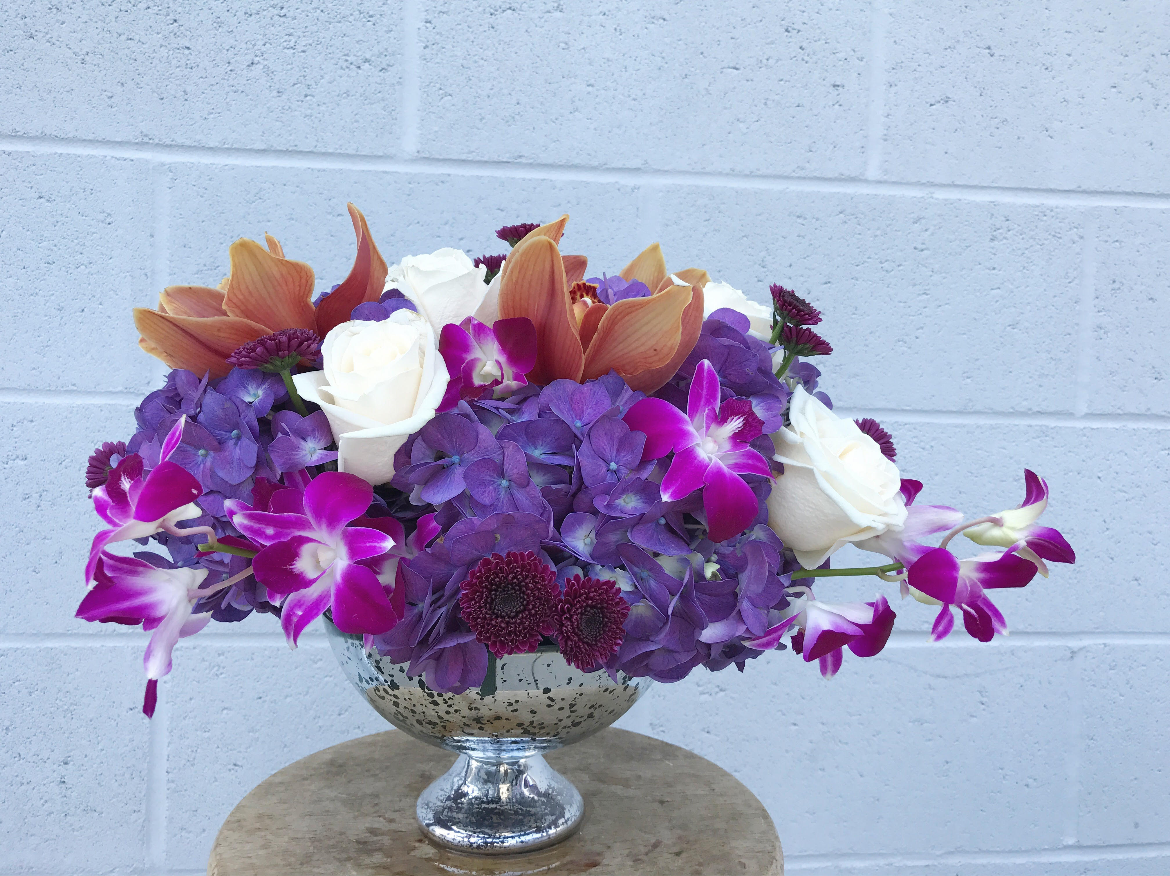 Purple hydrangeas, white roses, and orchids in a silver pedestal bowl