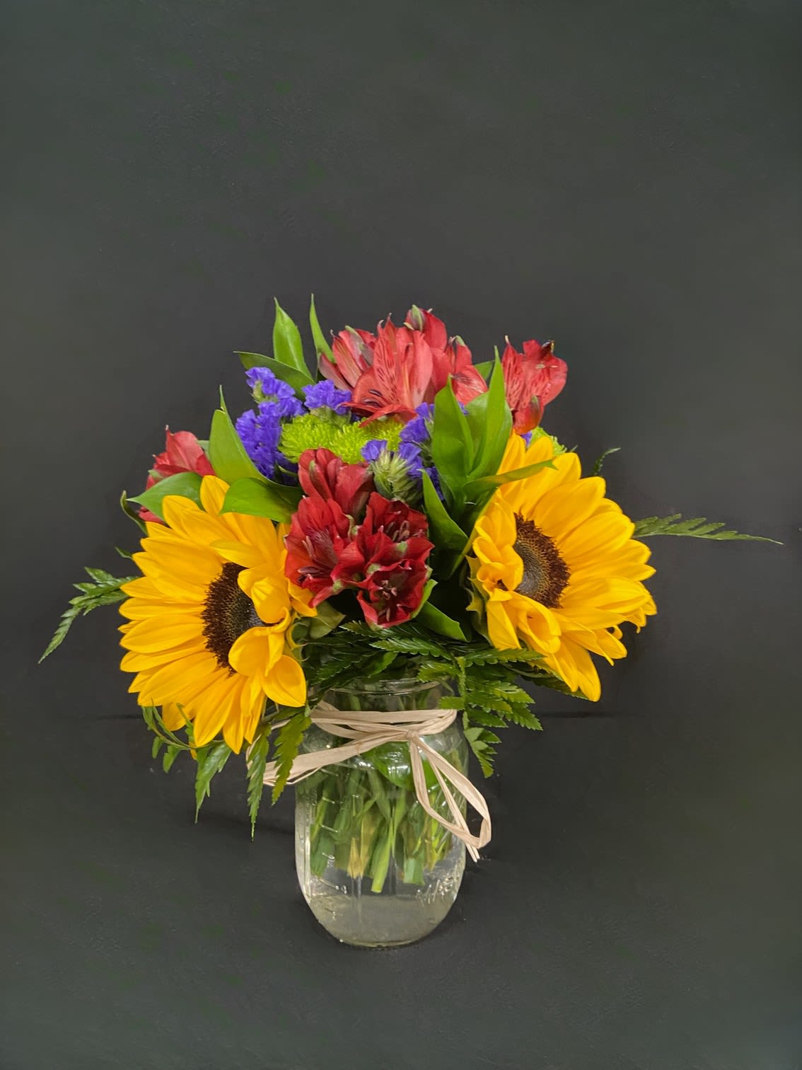 Summer Blooms Mason Jar - Colorful and vibrant - this mason jar is packed with gorgeous yellow sunflowers, red roses, red / pink / purple alstro, green CDN poms, and purple filler flowers.