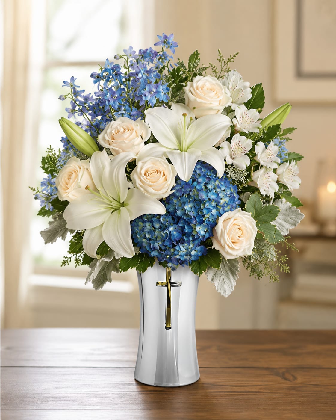 Vase of Remembrance  - A calm, reverent arrangement that offers a gentle, comforting presence for sympathy and funeral moments. This vase arrangement of lilies, roses and hydrangeas reads as a thoughtful memorial gift-the white lilies and creamy roses convey purity and remembrance while the blue hydrangea and delphinium bring soothing cool tones that calm the eye. The silver metallic vase with a cross  adds a subtle spiritual touch appropriate for religious services or quiet reflection at home.  The design pairs full, rounded clusters of blue hydrangea and creamy roses with upright white lilies and taller blue delphinium to create layered height and texture. Sprays of smaller white alstroemeria tuck between blooms for delicate detail. Finished in a flared silver vase with a cross on the front, the arrangement measures to fill a medium-to-large tabletop or memorial display and presents a serene color story of white, blue, and soft peach accents.  Send this as a meaningful way to express sympathy and support-the person you're thinking of will feel comforted and honored by its restrained elegance. Ordering and delivery notes: designed for immediate display with water-filled floral foam in the vase; replace water as needed and remove any wilted stems to extend freshness.