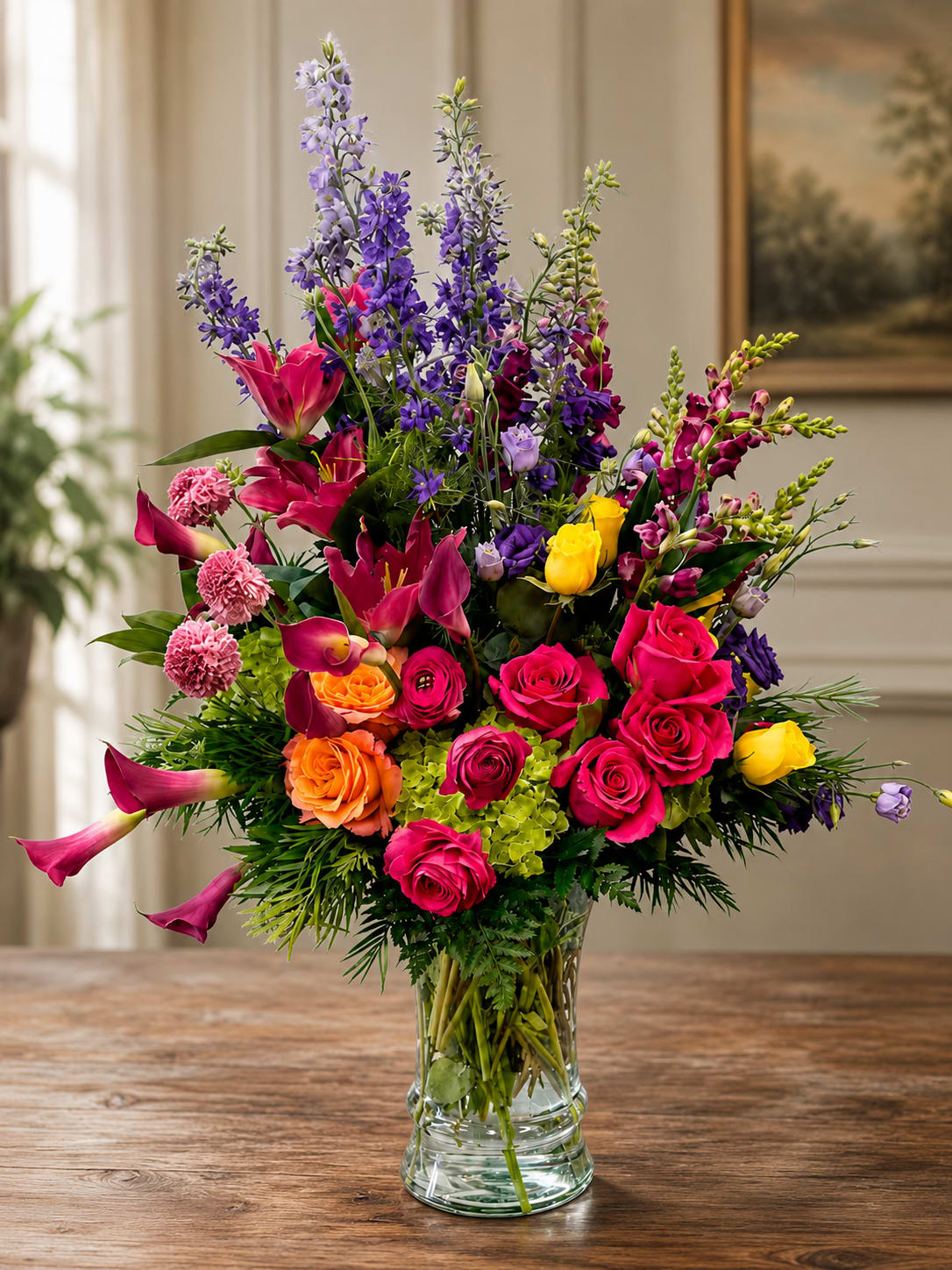 Vibrant Romance  - A bold cascade of color greets the eye - hot pink roses and deep burgundy mini calla lilies punctuated by bright yellow and orange blooms create the heart of this flower arrangement, perfect as an anniversary roses bouquet, a birthday surprise, or a vibrant Mother's Day gift. Spikes of purple and blue delphinium and snapdragons rise above the cluster to give the piece a tall, garden-like silhouette that reads both celebratory and romantic; it's the kind of arrangement that makes the person you're thinking of feel truly seen and adored.  Set in a clear glass vase, the design layers lime-green accent blooms and textural filler throughout for contrast and visual depth, with soft touches of lavender, peach and white balancing the saturated palette. The mix of roses, mini calla lilies, delphinium, snapdragons and hydrangea-like fullness gives varied texture and lasting presence on a mantel, dining table, or bedside. Ideal for occasions from graduations and housewarmings to get well wishes, sympathy, or just because moments, this arrangement also suits seasonal celebrations like Easter and springtime events while remaining a confident gift any day of the year. Care: refresh water every two days and trim stems slightly when re-cutting to extend vase life.