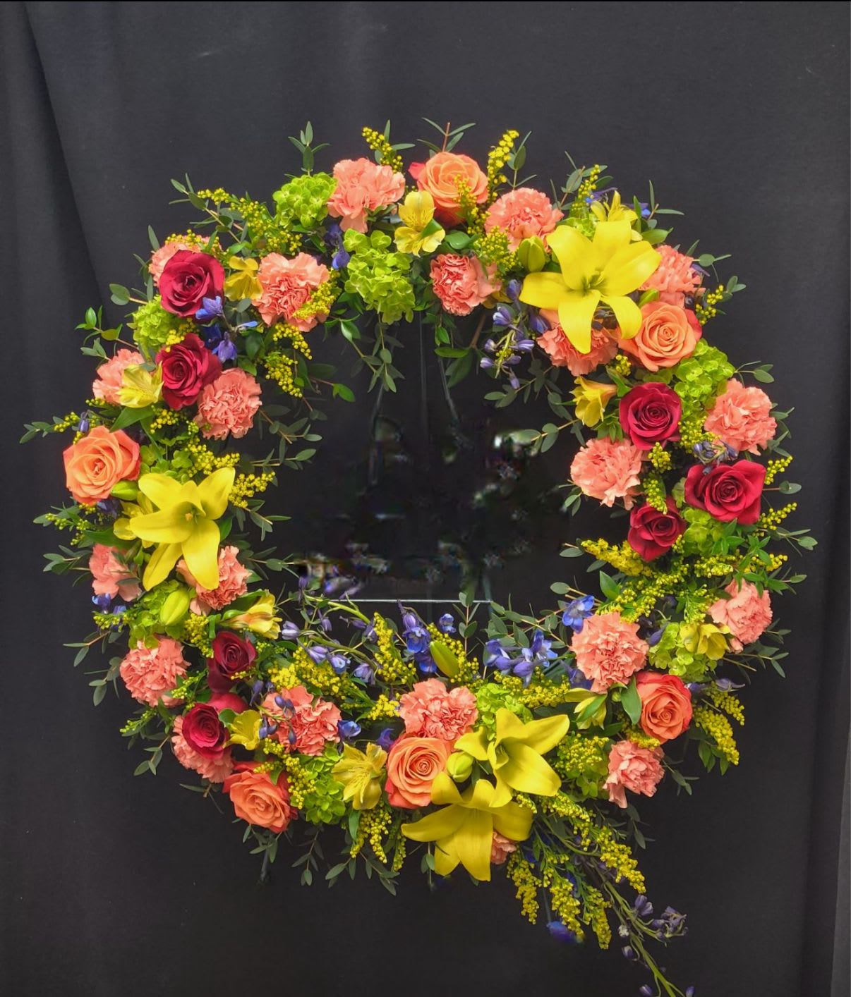 Circular floral wreath with roses, carnations, and lilies on a dark background
