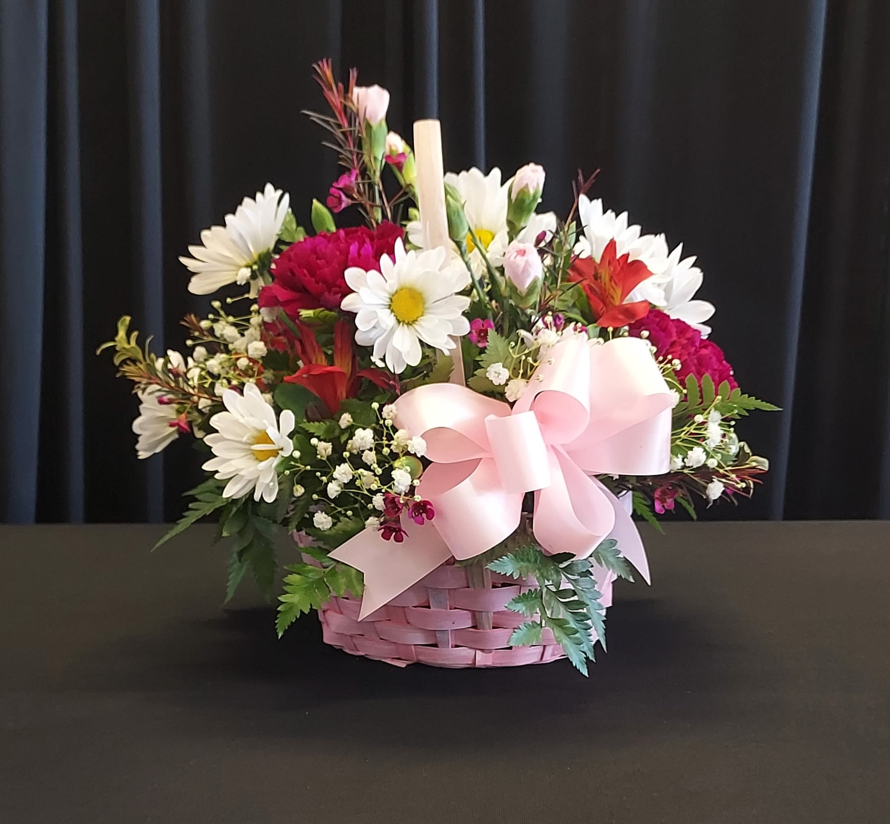A Basket for MOM - Bright and cheerful in a pink woven basket, this flower arrangement is a joyful choice for Mother's Day, a birthday, or just to say "I'm thinking of you." The mixed bouquet-presented in a charming basket with a large pale pink ribbon-pairs white daisy-like blooms with deep red and magenta carnations (and other seasonal flowers) to create a warm, celebratory look that will make Mom feel truly seen.  Textured sprigs and small filler flowers give the arrangement a full, garden-style silhouette with lively contrasts of white, pink, red, and green. The woven basket and soft ribbon make this a ready-to-display gift that looks polished on a kitchen table or bedside dresser; included candles add a cozy, gift-ready touch. Perfect for mothers who love classic florals with a playful pop of color, the size and materials make it easy to place and enjoy right away-no vase needed.