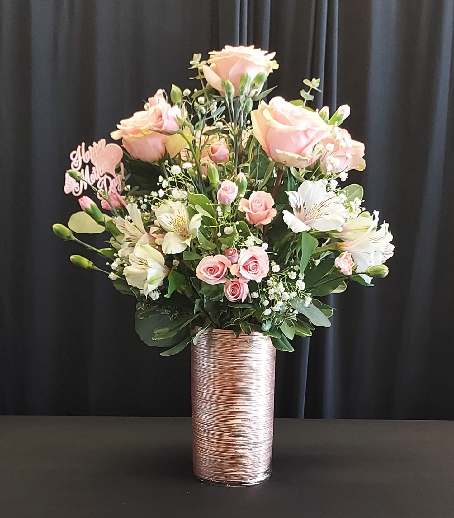 I Love Pink  - Soft pink roses and delicate spray roses mingle with white alstroemeria in a romantic, feminine flower arrangement perfect for a birthday bouquet, anniversary roses alternative, or a heartfelt thank-you gift. The soft, pale palette and polished presentation make this arrangement feel both celebratory and intimate-the kind of gift that makes her feel truly seen and cherished.  Arranged in a tall metallic rose-gold cylindrical vase, the design is full and rounded with layered blossoms and airy filler accents for gentle movement and texture. Expect a mix of garden-style roses, smaller spray roses, and crisp white alstroemeria framed by fresh greenery; a small pink decorative pick tucked into the left side adds a sweet, personalized touch. The height of the vase gives the bouquet elegant presence on a mantel or dining table and keeps stems neatly supported for lasting enjoyment.  Ideal for the person you want to celebrate-mothers, partners, or friends-this arrangement sends a message of affection and appreciation without being over-the-top. For best enjoyment, keep in fresh water, out of direct sunlight, and trim stems slightly if needed when you refresh the vase; arrangements arrive ready to display and make an immediate, lovely impression.