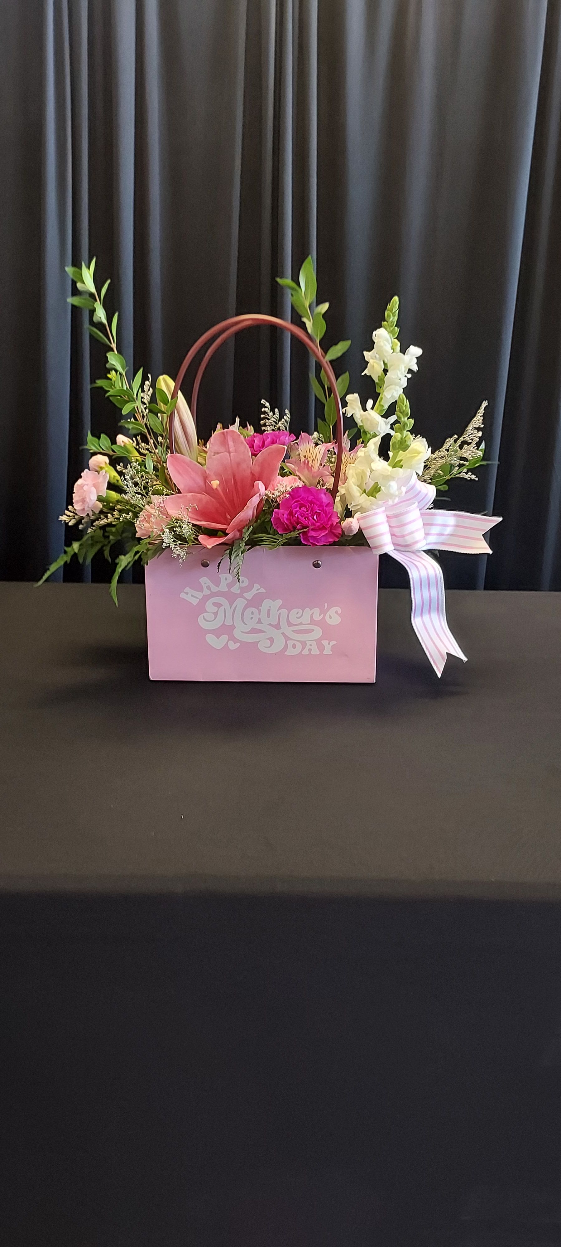 Mommy's Purse  - A bright pink lily takes center stage in this feminine Mother's Day flower arrangement, styled like a charming mommy's purse that reads as a heartfelt birthday, thank-you, or spring gift. The flower arrangement and occasion (Mother's Day, birthday, thank-you) are woven into the design so the person you're celebrating will feel seen and adored the moment it arrives.  The arrangement mixes vivid pink carnations, pale alstroemeria, and tall white snapdragon spikes with airy filler and green accents for layered texture and height. Presented either in a pink handled basket with an arched handle and a Mother's Day message or in a coordinating pink gift box, it's finished with a pink-and-white striped ribbon and decorative bow for a polished, gift-ready look.  Perfect for moms, grandmothers, or a special woman in your life, this purse-style bouquet reads as both festive and personal - fresh, fragrant, and full of spring color. Available in a compact, hand-arranged size that's easy to display on a table or countertop; enjoy the blooms in fresh water and trim stems as needed to extend vase life.