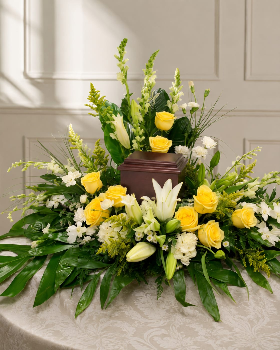 Yellow and White Cremation Wreath  - A low, elongated spray of yellow and white roses and lilies designed for sympathy and funeral settings that still feels quietly uplifting. The horizontal, triangular shape tapers gently at both ends so the arrangement creates a graceful, linear profile on a memorial table - the kind of tribute that helps the person you're honoring feel seen and remembered.  Clusters of bright yellow roses sit beside open and budding white lilies, with other complementary blooms and bold green leaves fanning beneath to add structure and length. The palette of sunny yellow, pure white, and fresh green reads calm and hopeful; the overall size and horizontal format make it ideal as a centerpiece on a low table.  Made to comfort and celebrate a life, this wreath sends a thoughtful message of remembrance and warmth for funeral services or sympathy visits. Arrange for delivery to the service location and keep stems in clean water until placement; if you need a different size or personalization, let us know and we'll help create the right tribute.