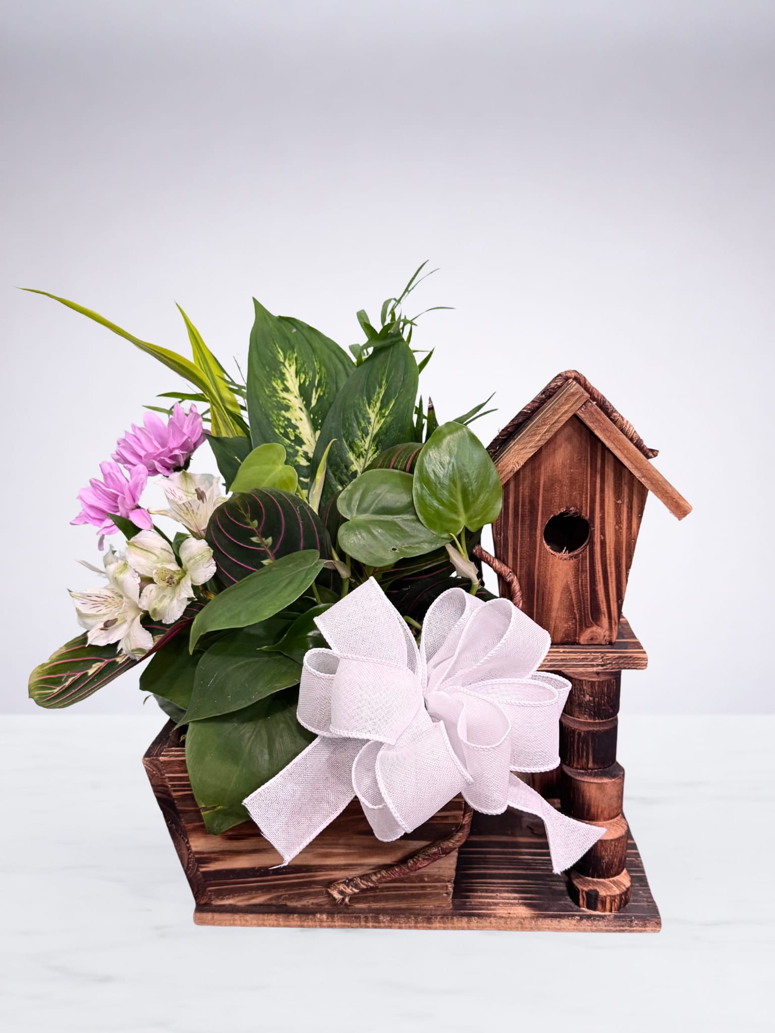 Lush green plants with pink and white flowers in a rustic wooden birdhouse planter with a white bow