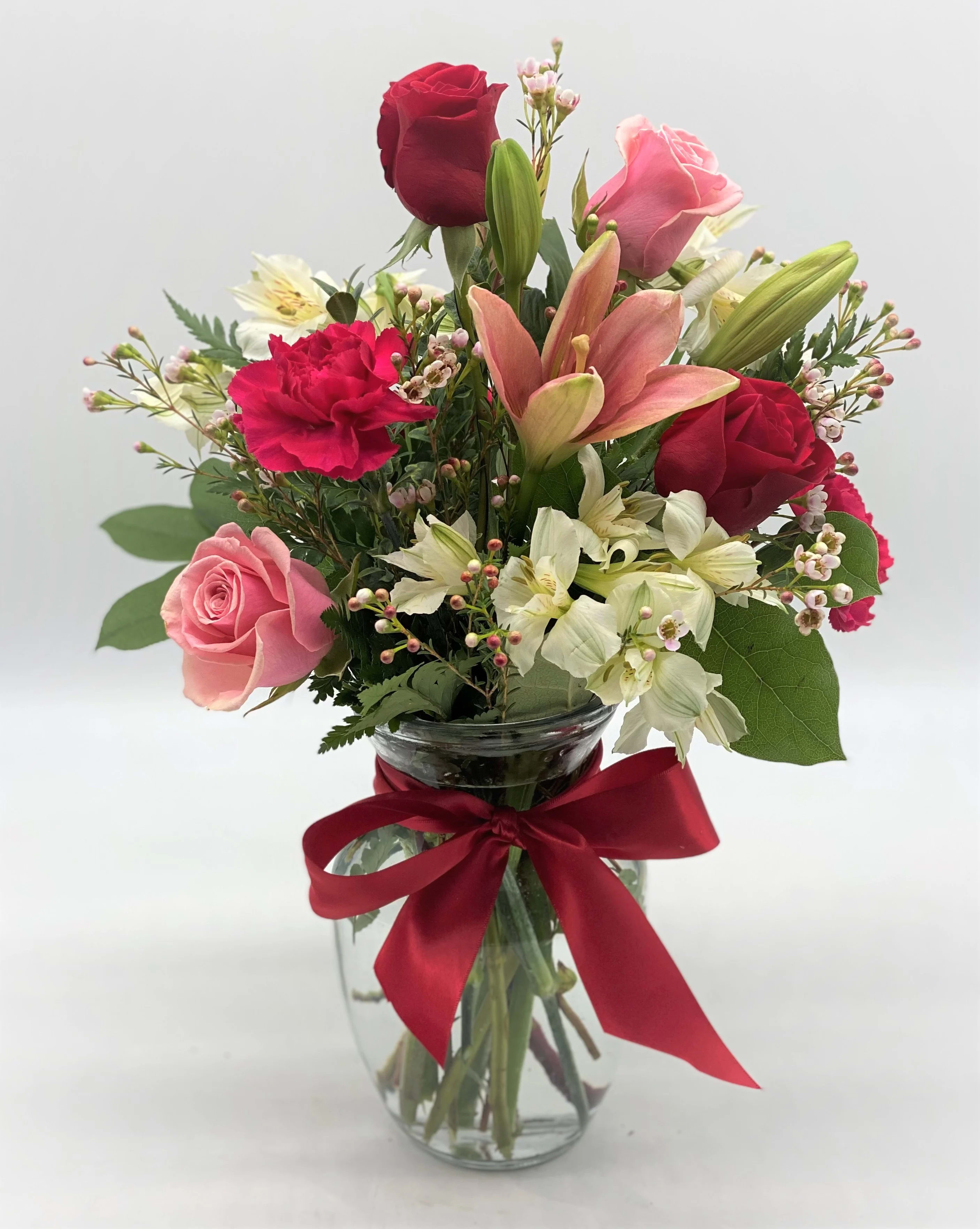 Mixed bouquet of roses, lilies, and carnations in a glass vase with a red ribbon