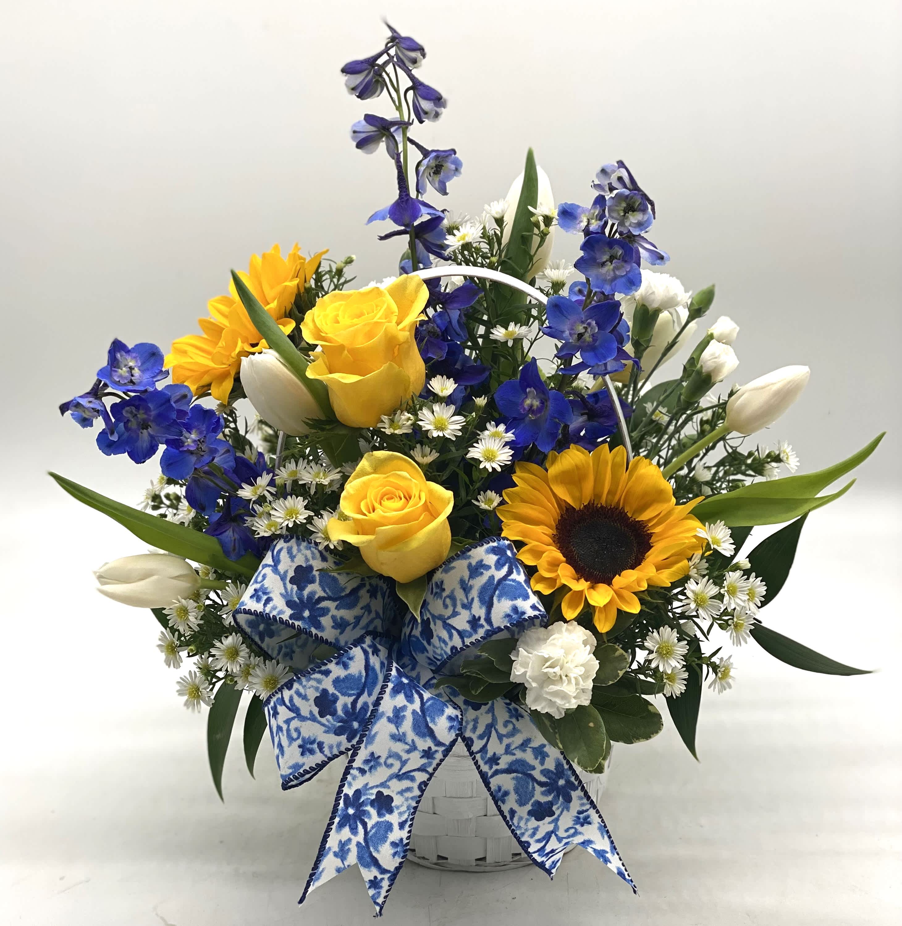 Basket arrangement with yellow roses, sunflowers, blue flowers, and white blooms