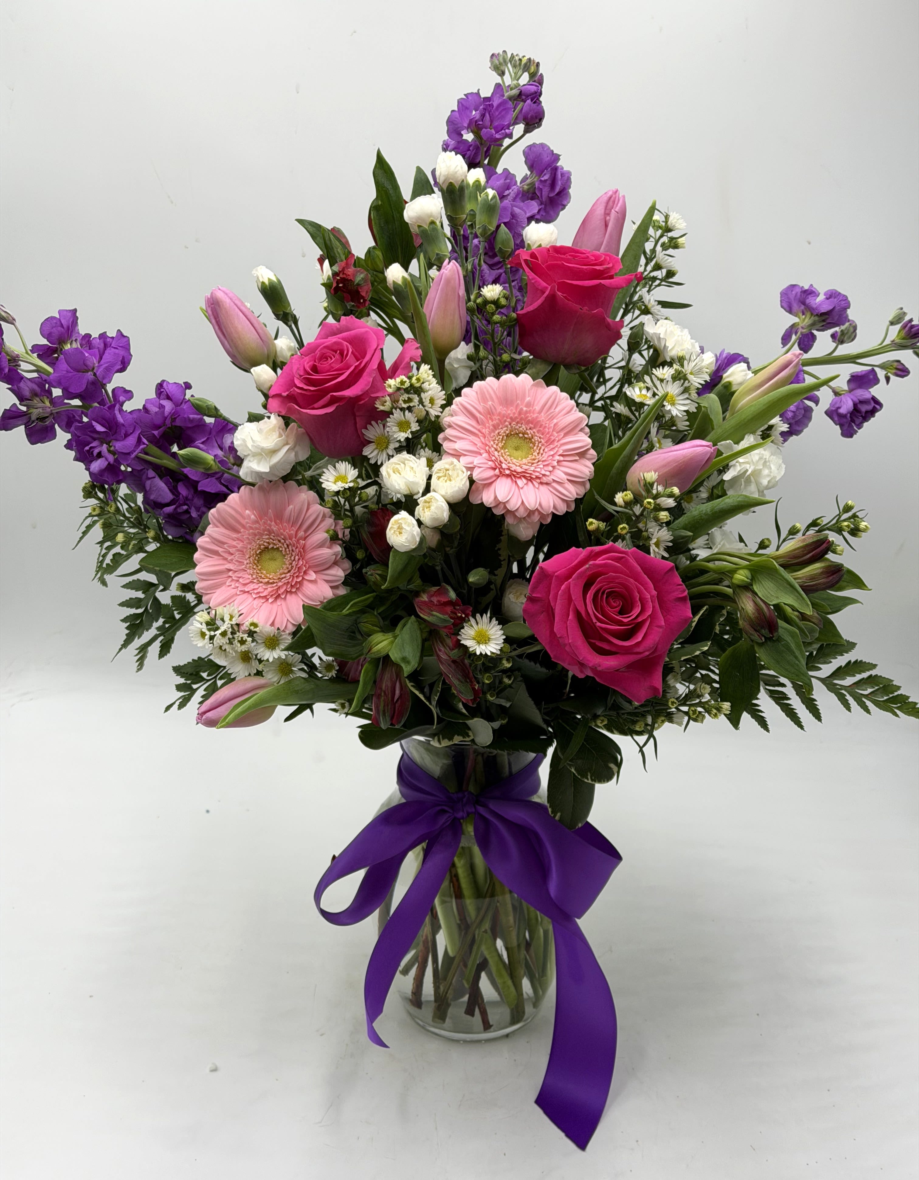 Mixed bouquet of pink and purple flowers in a glass vase with a purple ribbon