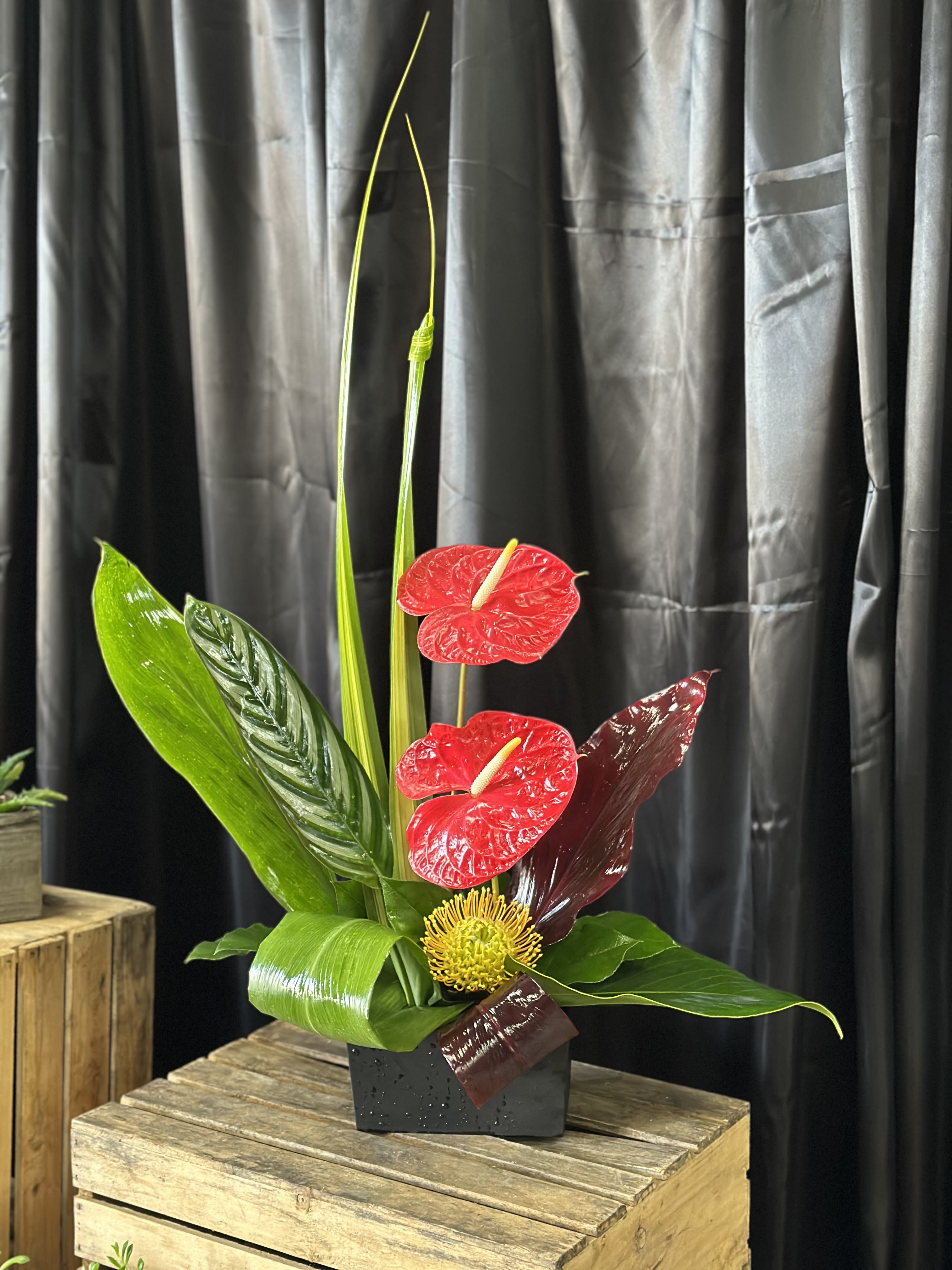 Tropical arrangement with red anthuriums and a yellow pincushion bloom in a black vase