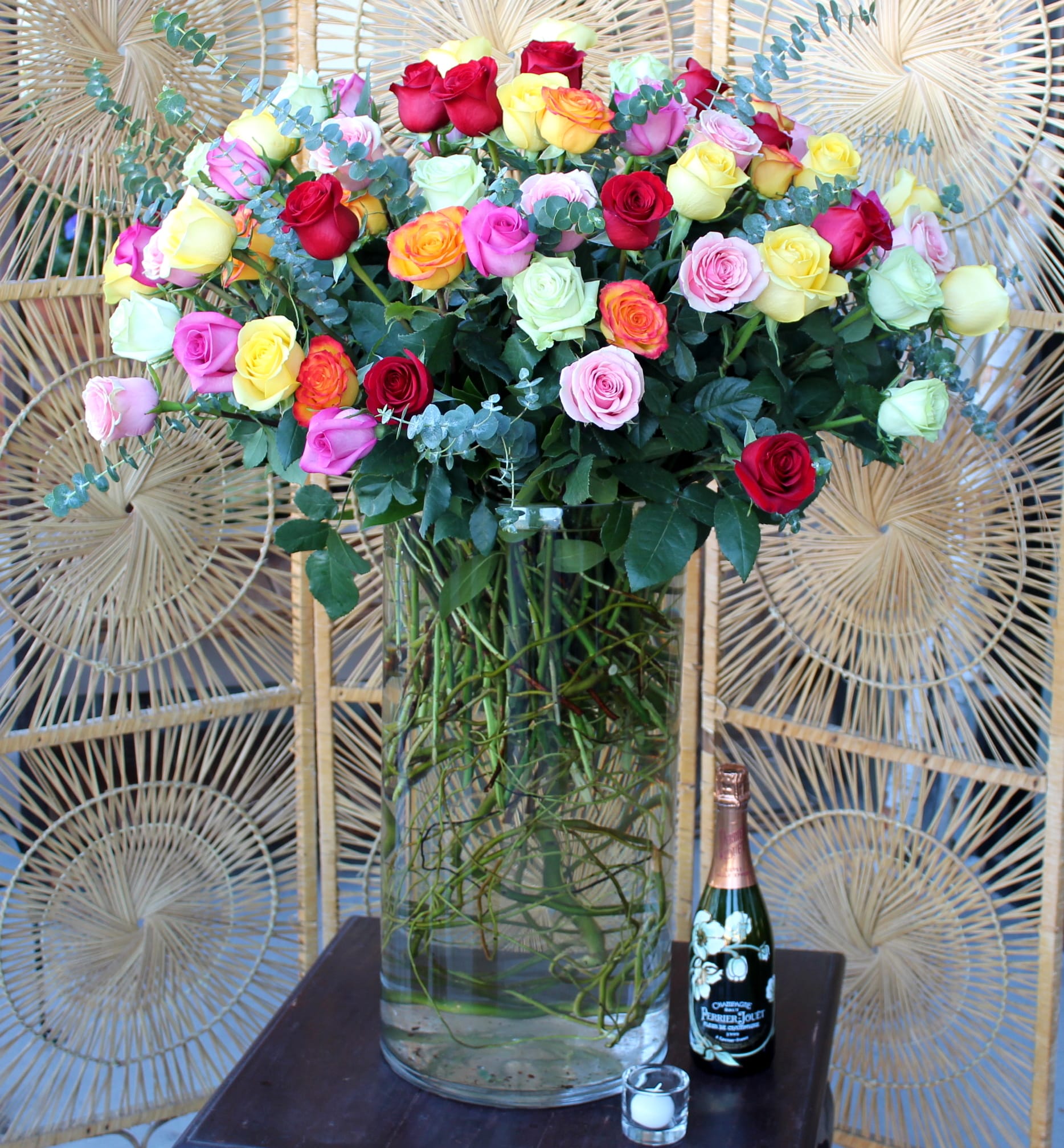 Mixed roses in a tall glass vase with a champagne bottle and candle